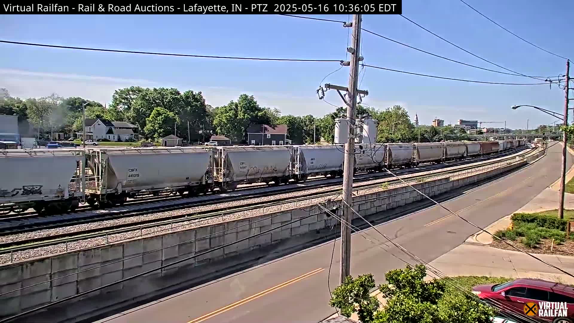 A long freight train of light gray boxcars travels along railroad tracks next to a road on a sunny day.