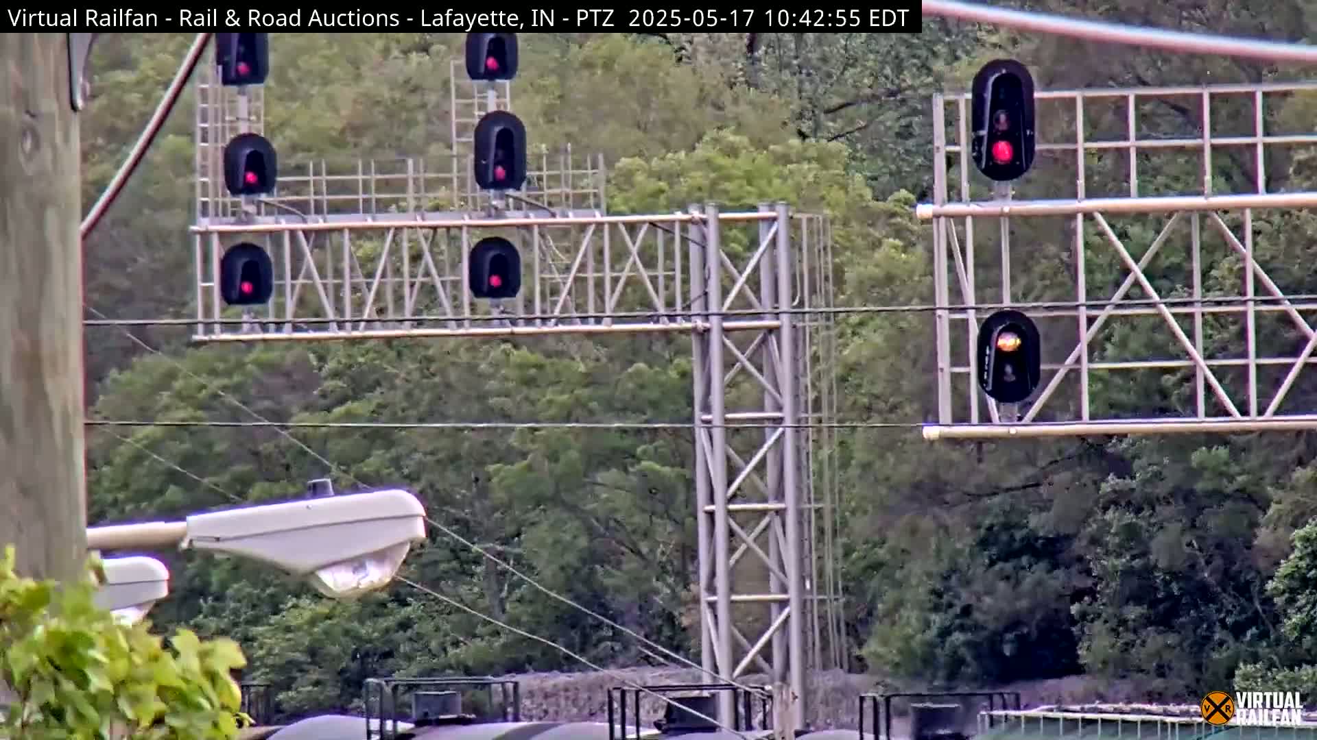 A metal framework with multiple red railroad signal lights is visible against a backdrop of green trees in daytime.