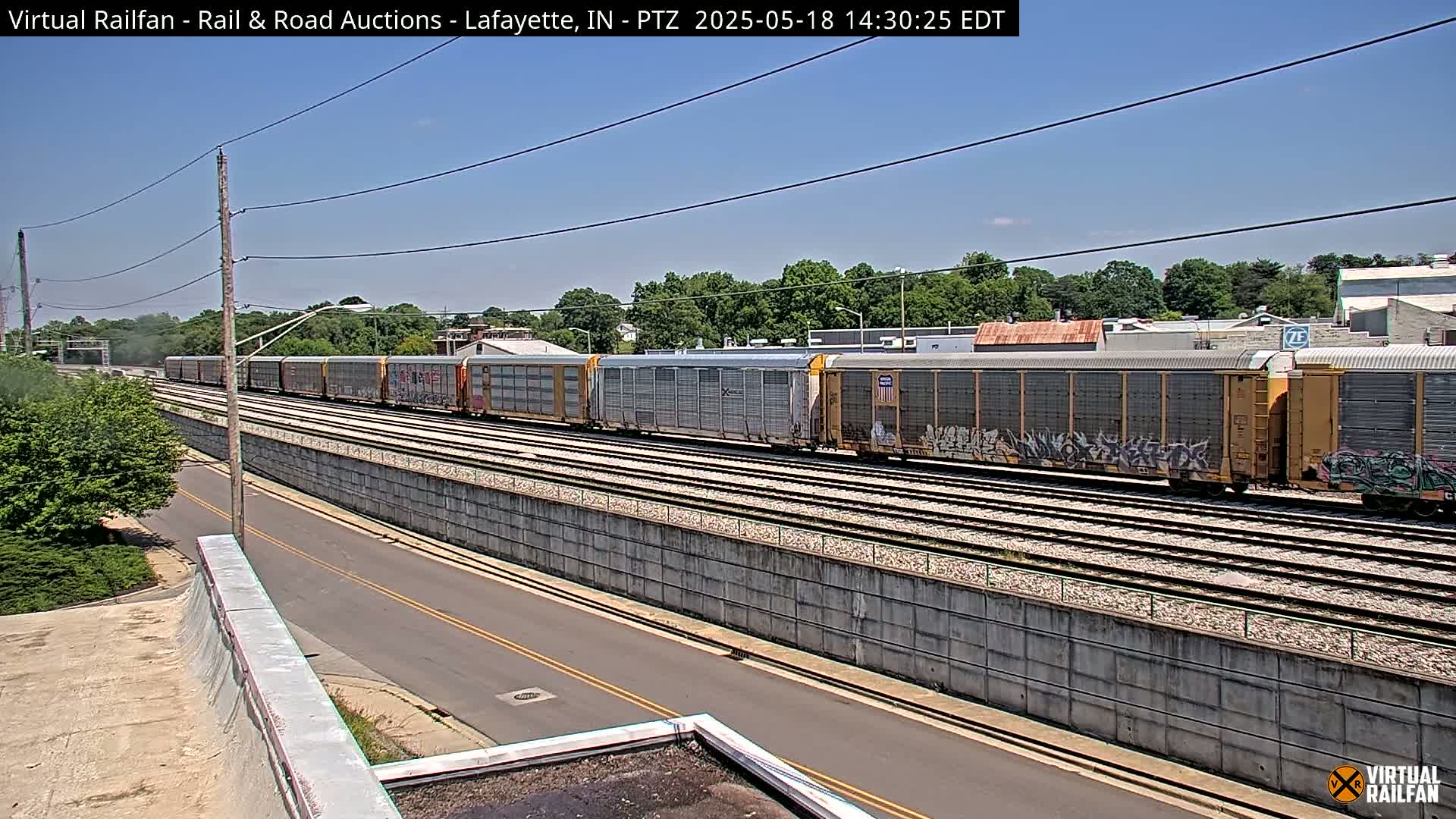 A long freight train with graffiti-covered cars passes along railroad tracks beside a road on a sunny day.