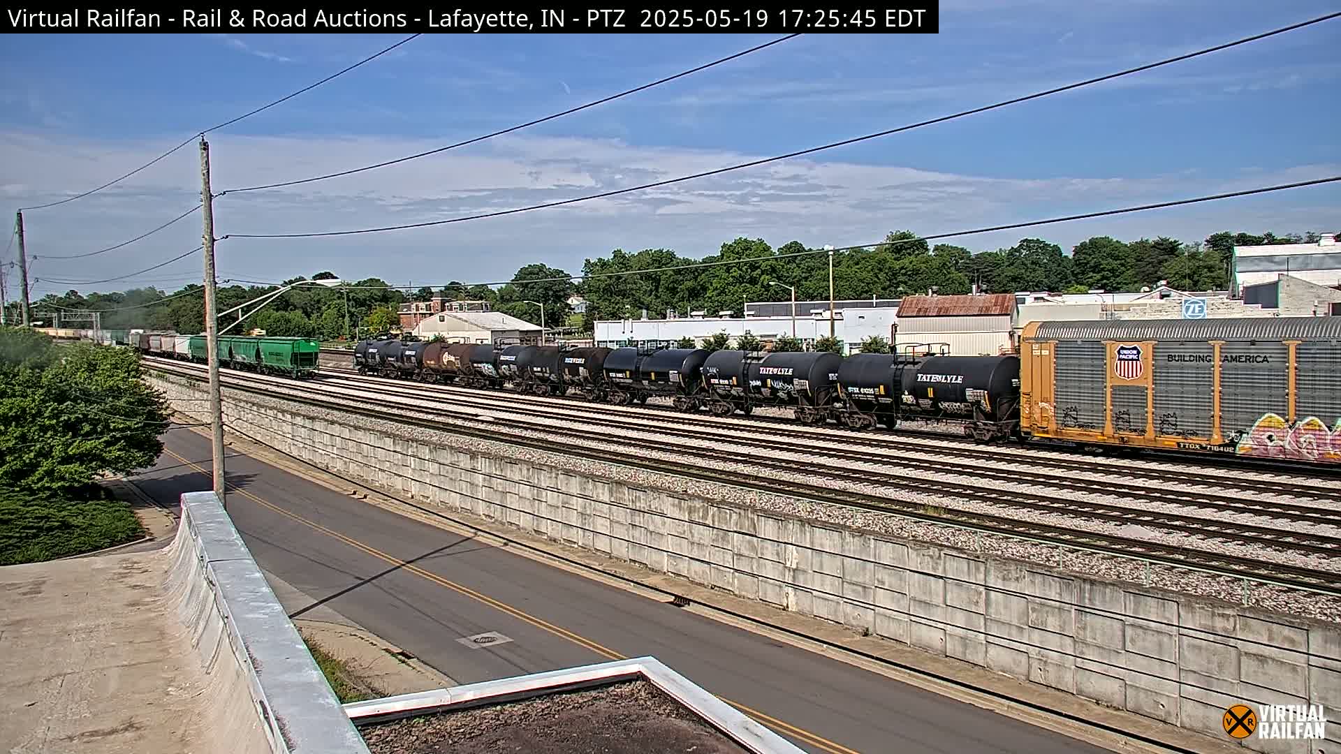 A long freight train, including tanker cars and boxcars, passes by on a sunny day along a set of railroad tracks beside a road and a concrete retaining wall.