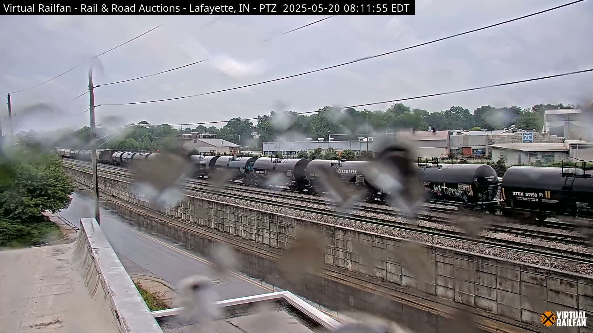 A long freight train of tanker cars passes by on a rainy day, viewed from an elevated position overlooking a rain-slicked road and railway embankment.