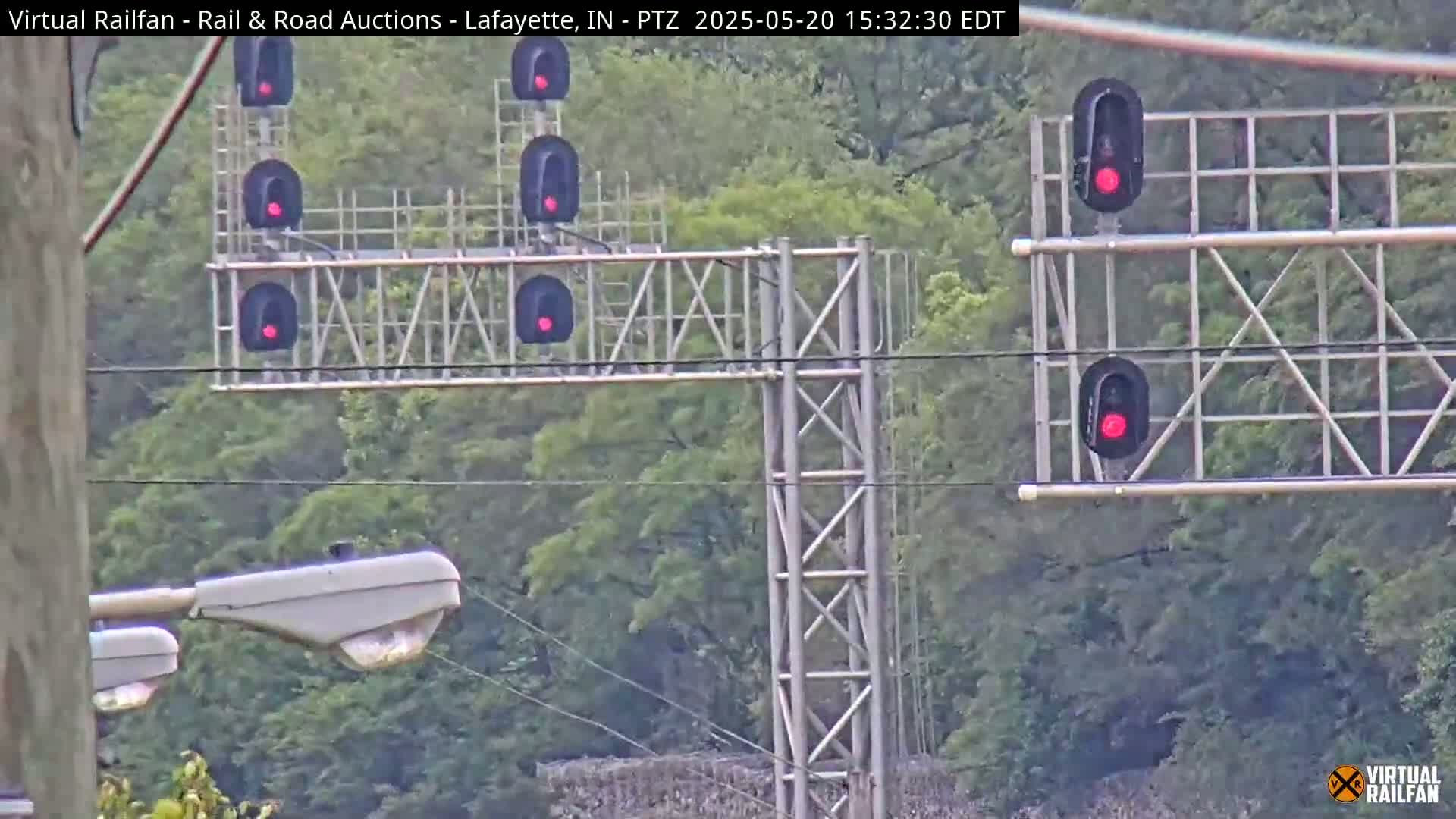 A metal railway signal structure with multiple red lights is situated in front of a lush green forest on an overcast day.