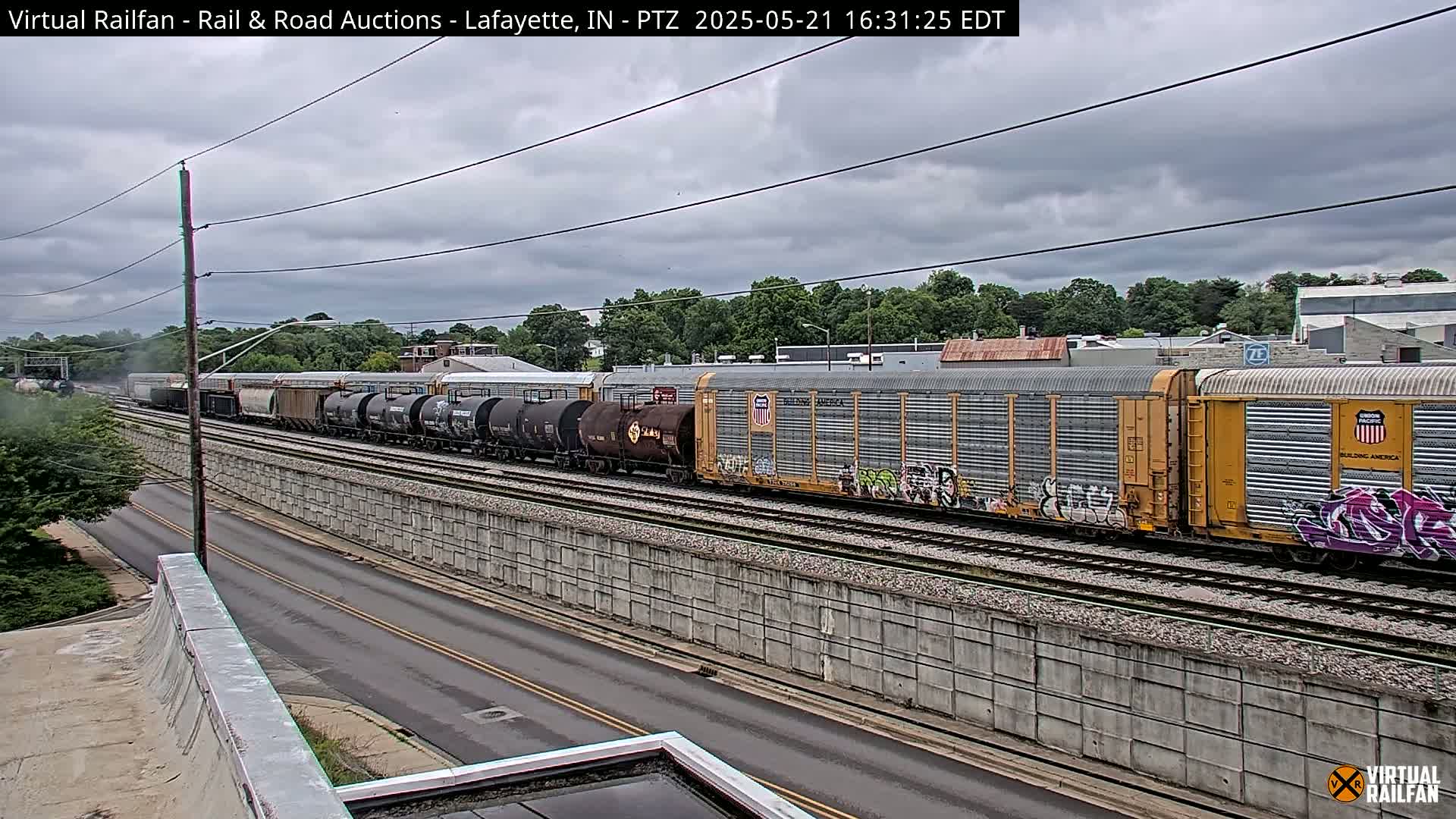 A long freight train passes under an overcast sky beside a road and a concrete retaining wall.