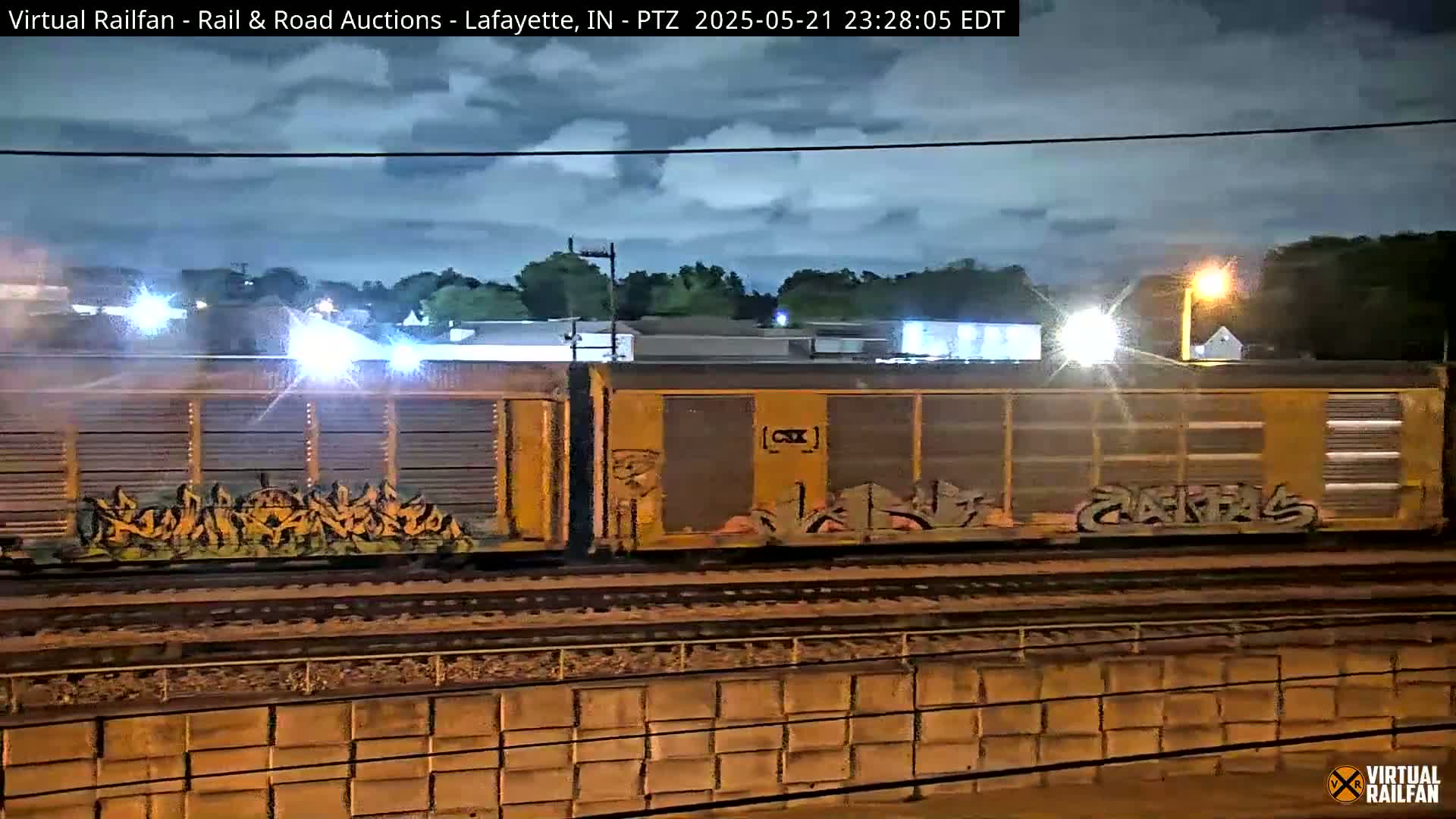 Under a partly cloudy night sky, a train with graffiti-covered cars is seen moving along the tracks near a low wall.