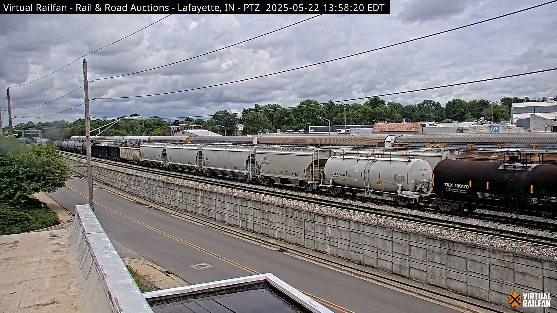 A long freight train passes under an overcast sky along a road next to a concrete wall.