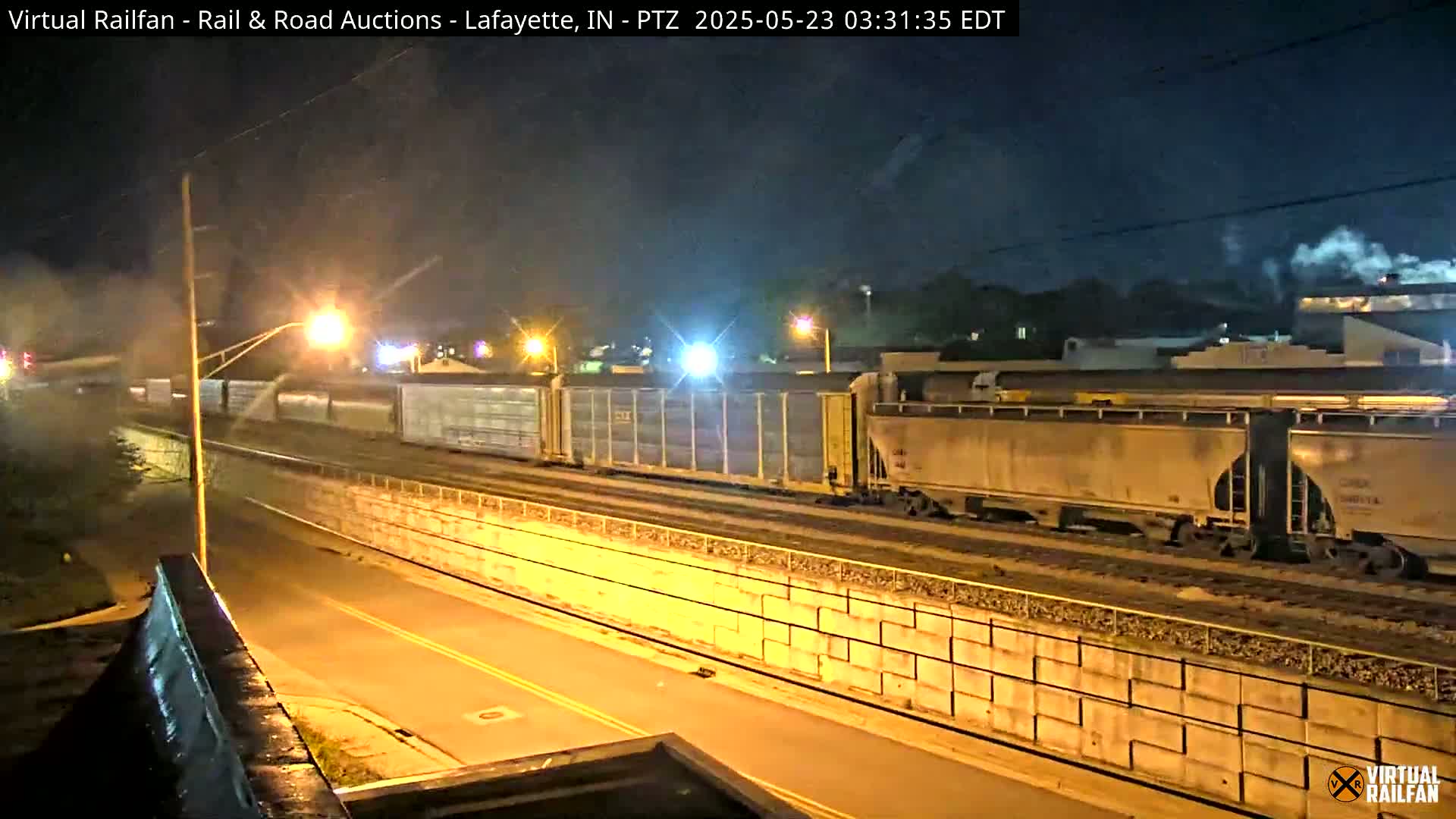 At night, a long freight train passes under a street illuminated by streetlights, beside a high retaining wall.