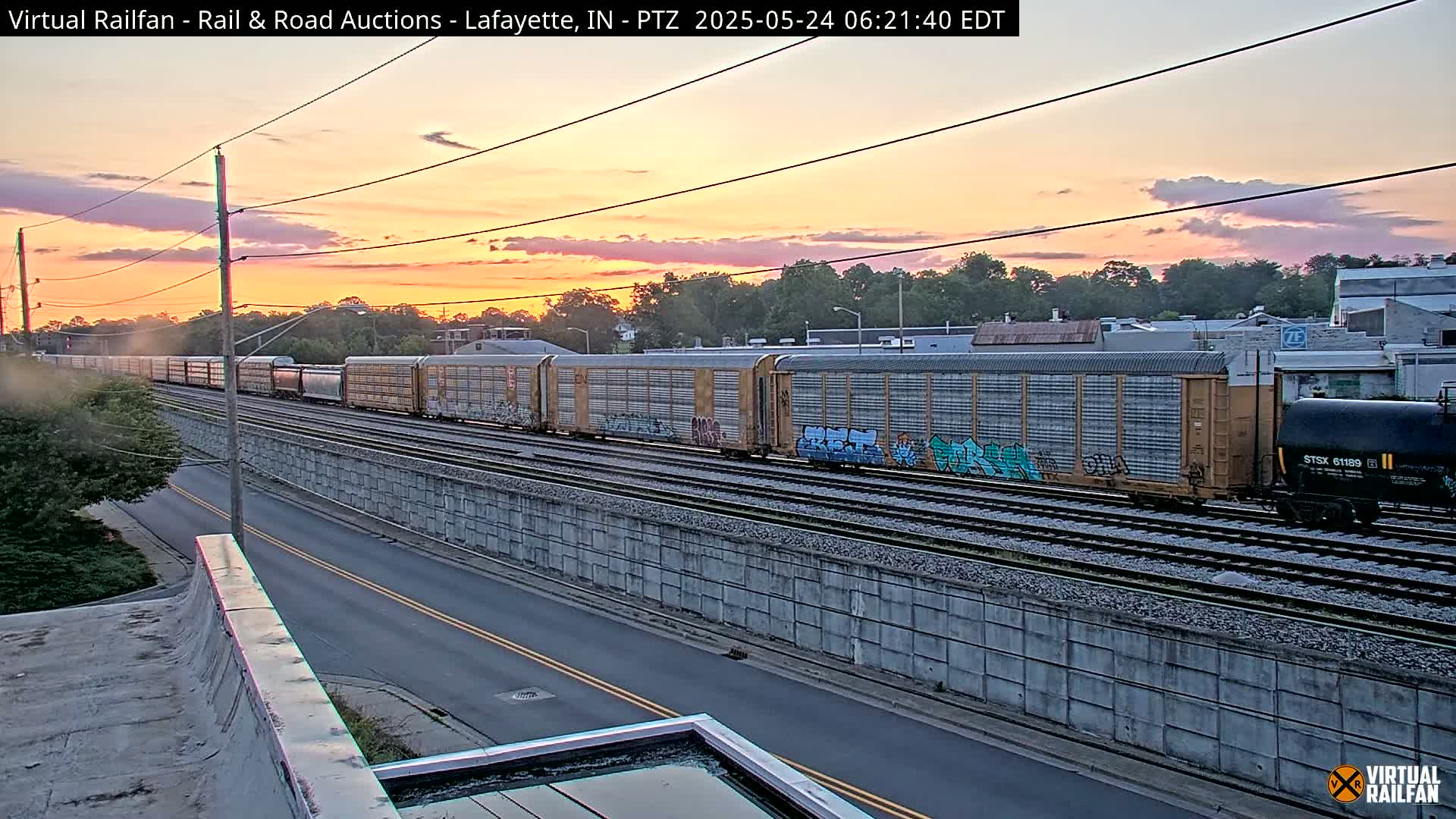 A long freight train with graffiti-covered cars passes under a sunset sky next to a road.