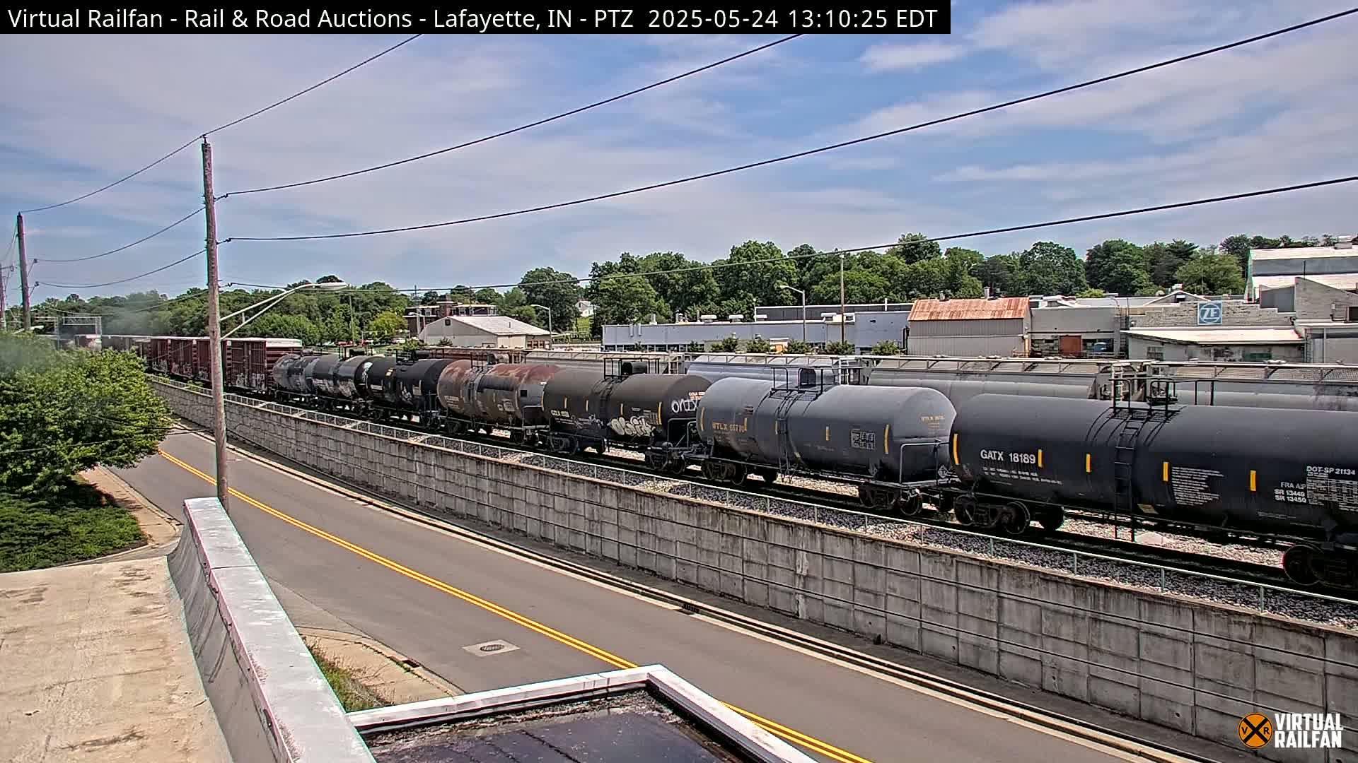 A long train of tanker cars is parked next to a road on a partly sunny day.