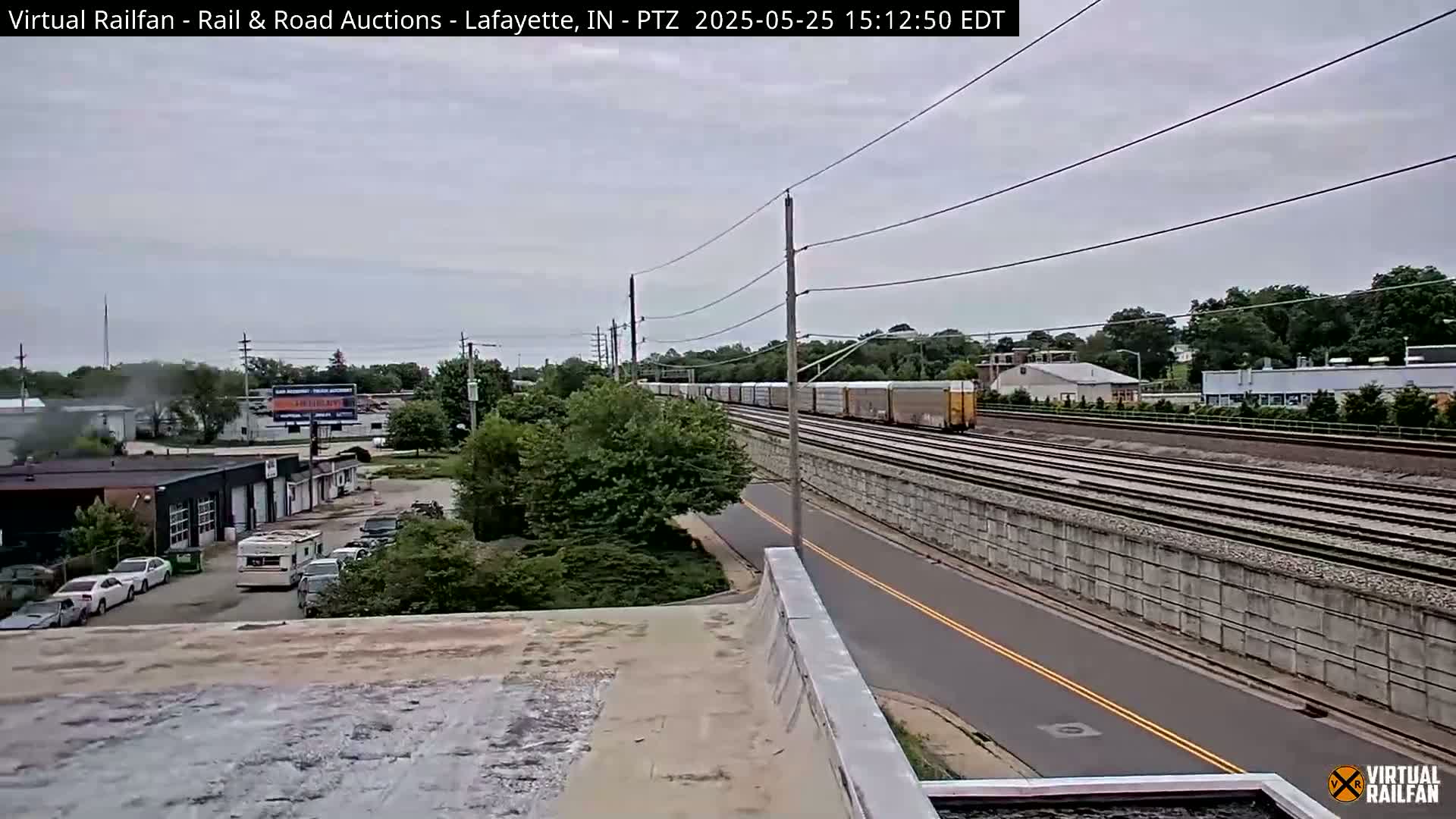 A long freight train passes by under an overcast sky, next to a road and a low wall separating the road and railway.