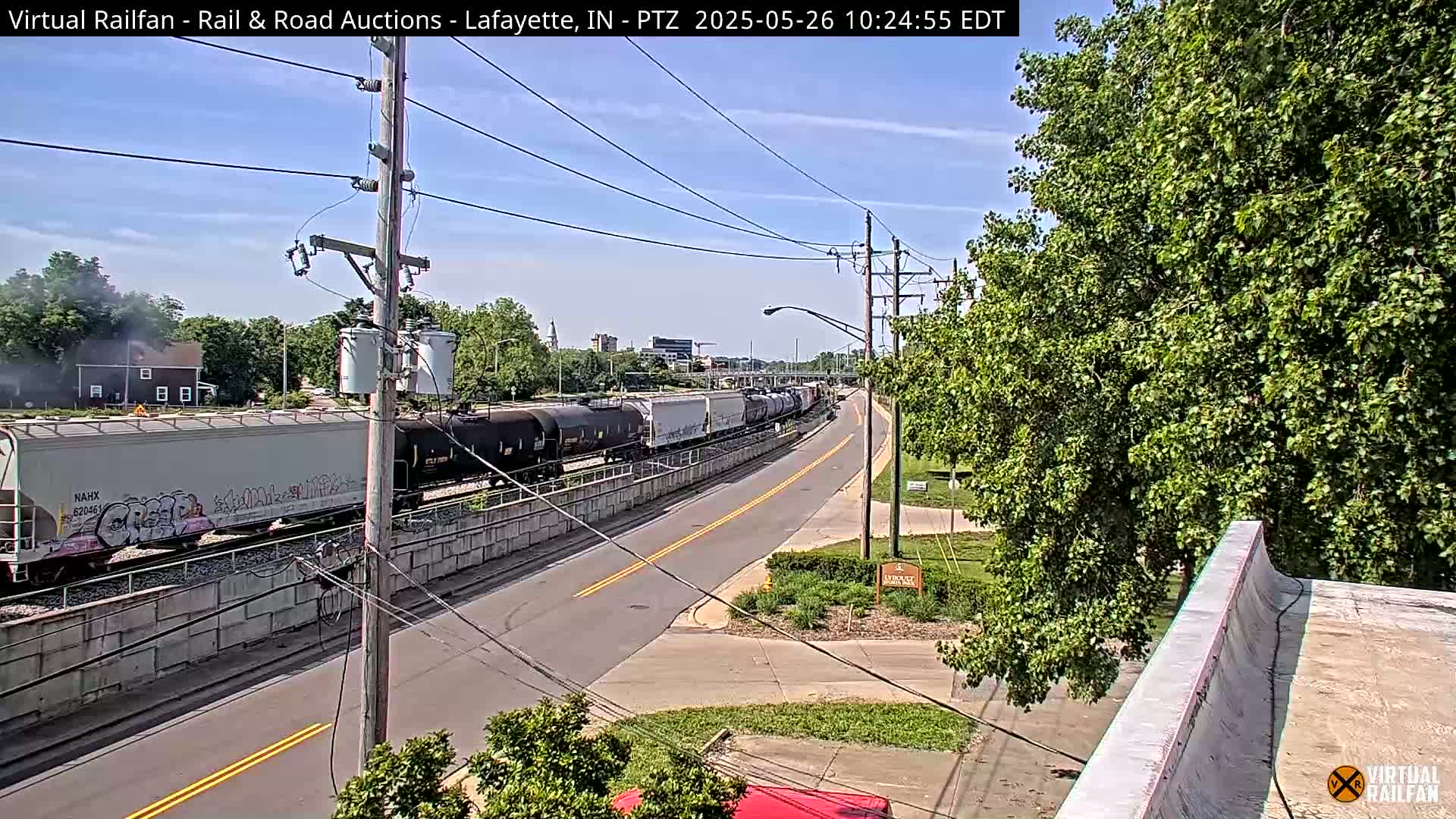 A long freight train passes by a road on a sunny day, with green trees lining the roadside.