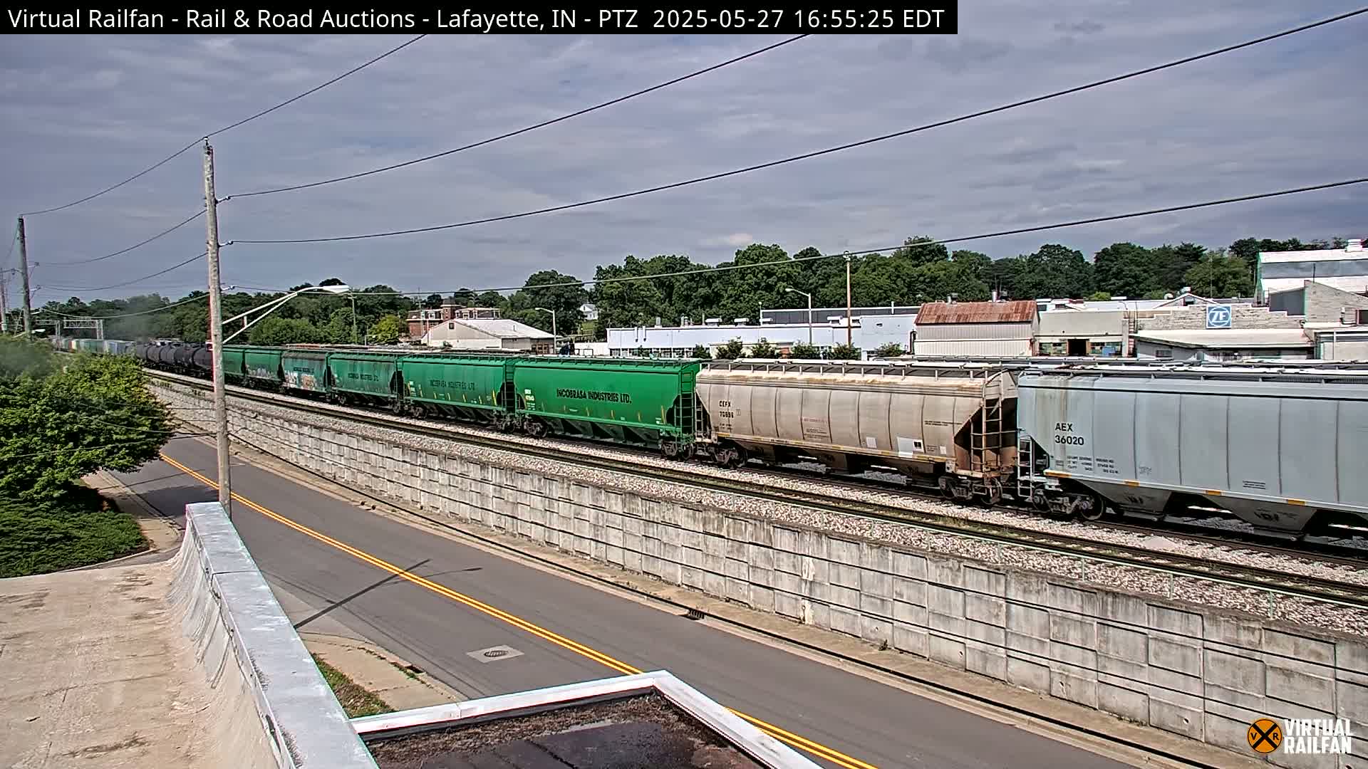 A long freight train, including green and gray cars, passes by under a partly cloudy sky.