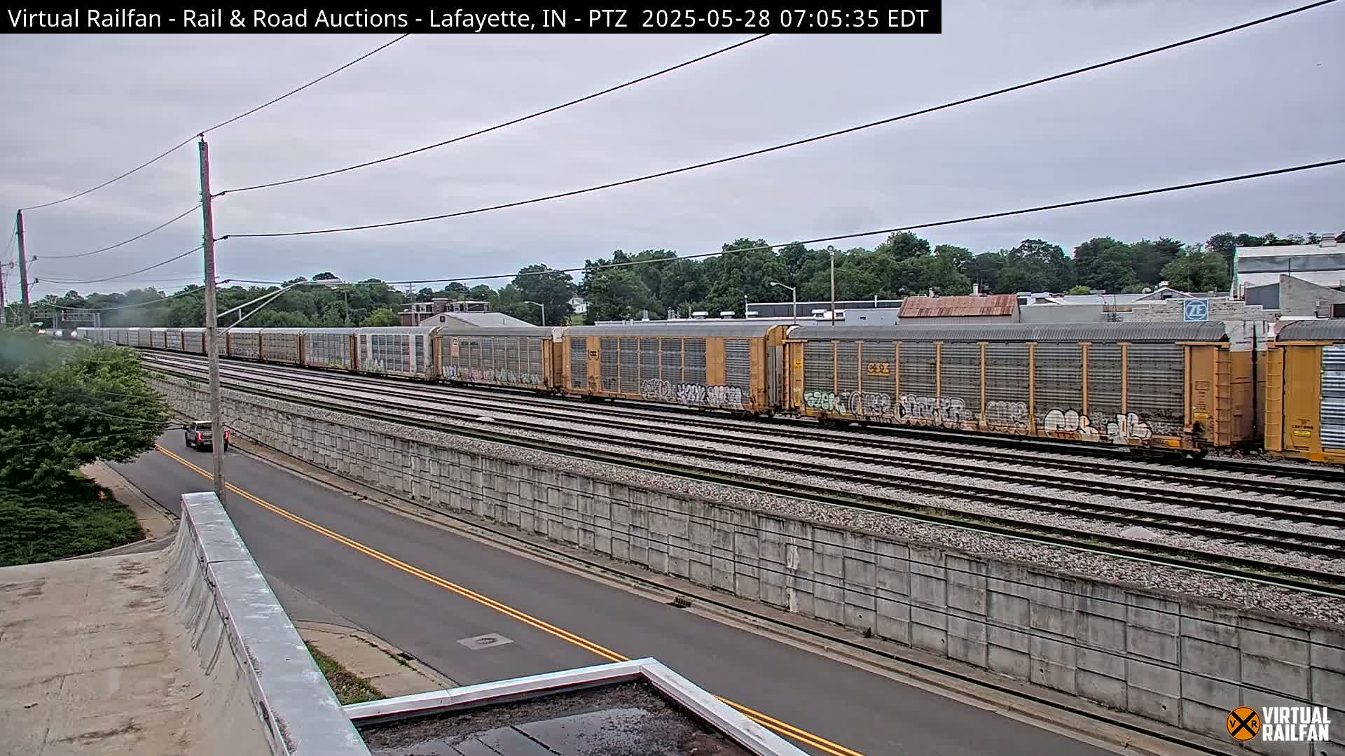 A long freight train with graffiti-covered cars passes by a road under an overcast sky.