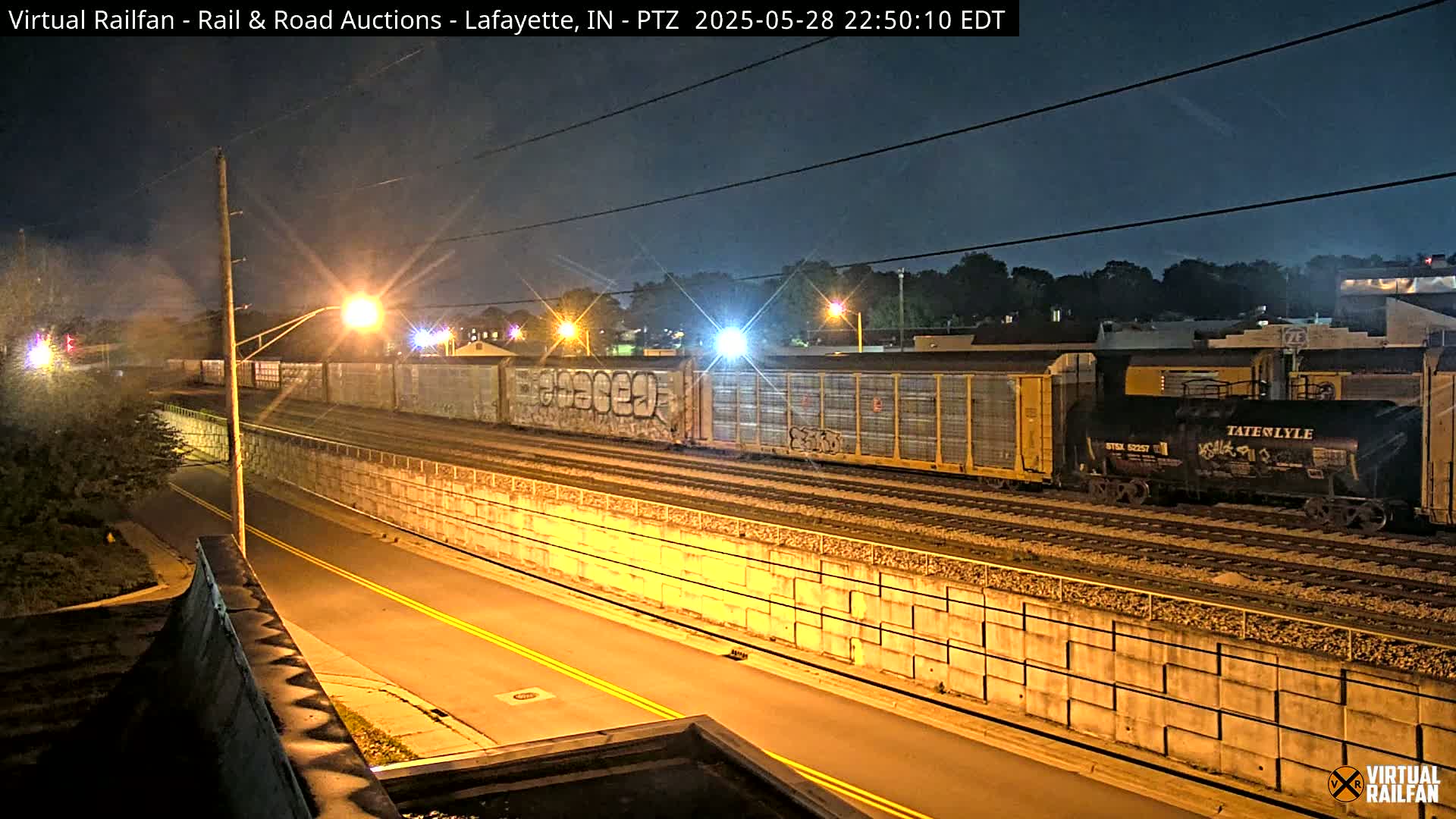 At night, a freight train with graffiti-covered cars sits on tracks beside a roadway bordered by a concrete wall.