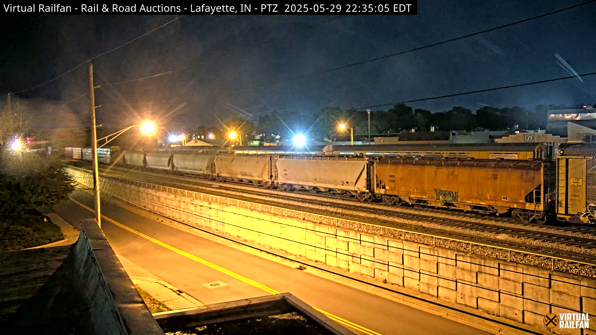 A long freight train is illuminated by streetlights at night, next to a concrete wall and a road.