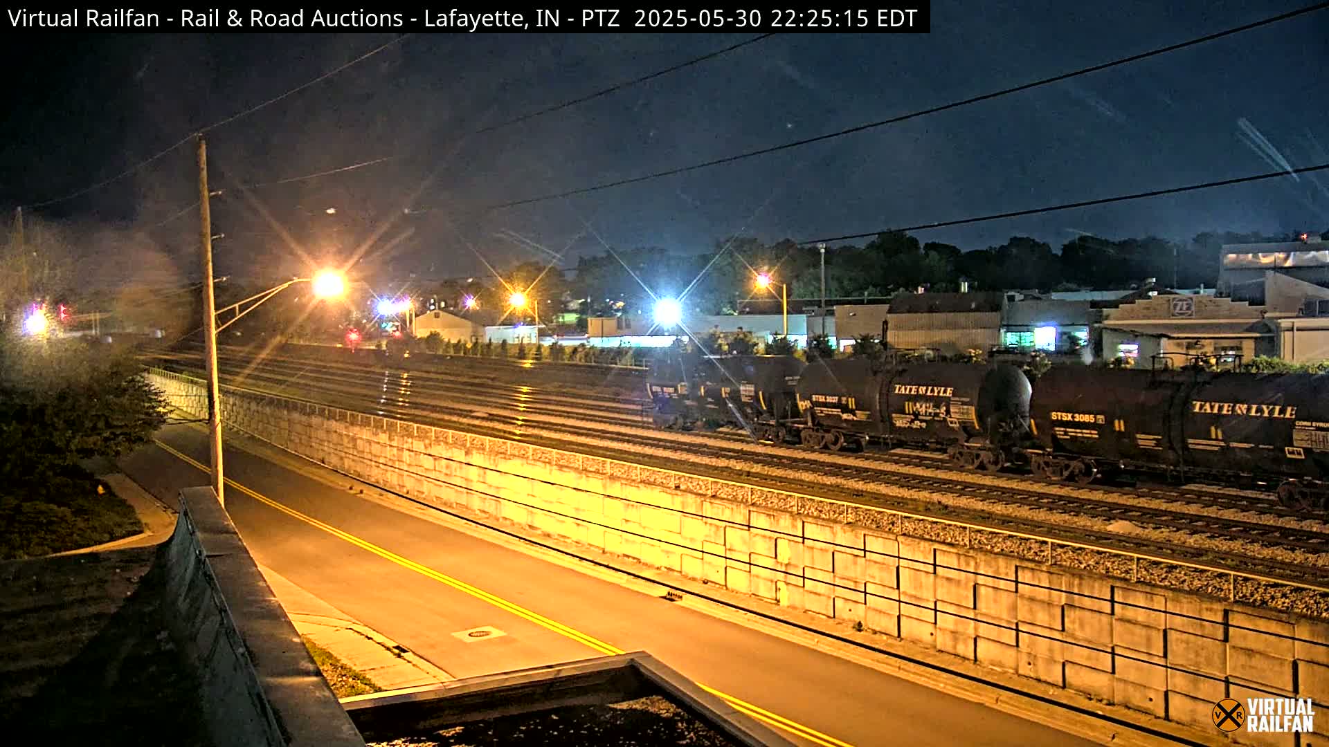 At night, a line of dark rail cars sits on tracks beside a brightly lit road separated by a concrete wall.