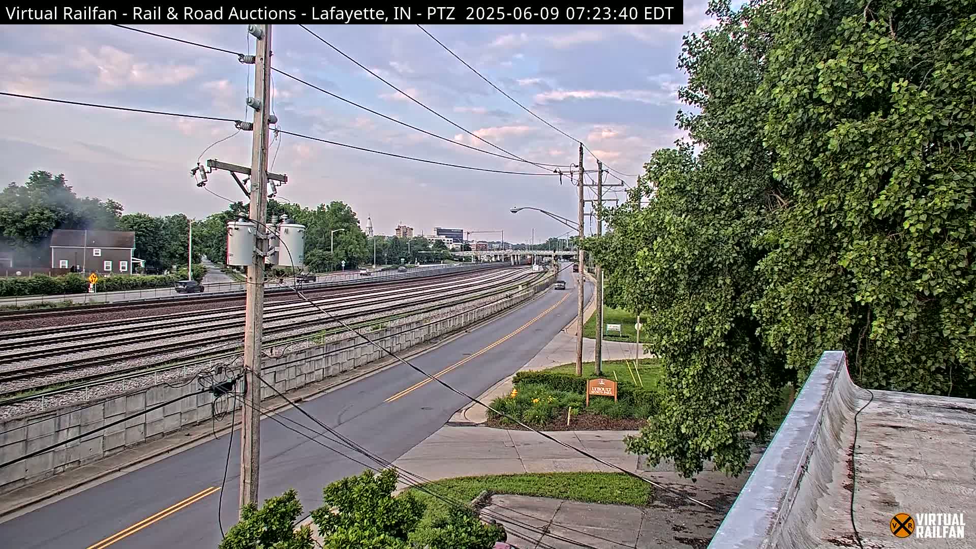 A partly cloudy sky overlooks a road and parallel railway tracks, with lush green trees lining the road and a concrete wall separating the road from the tracks.