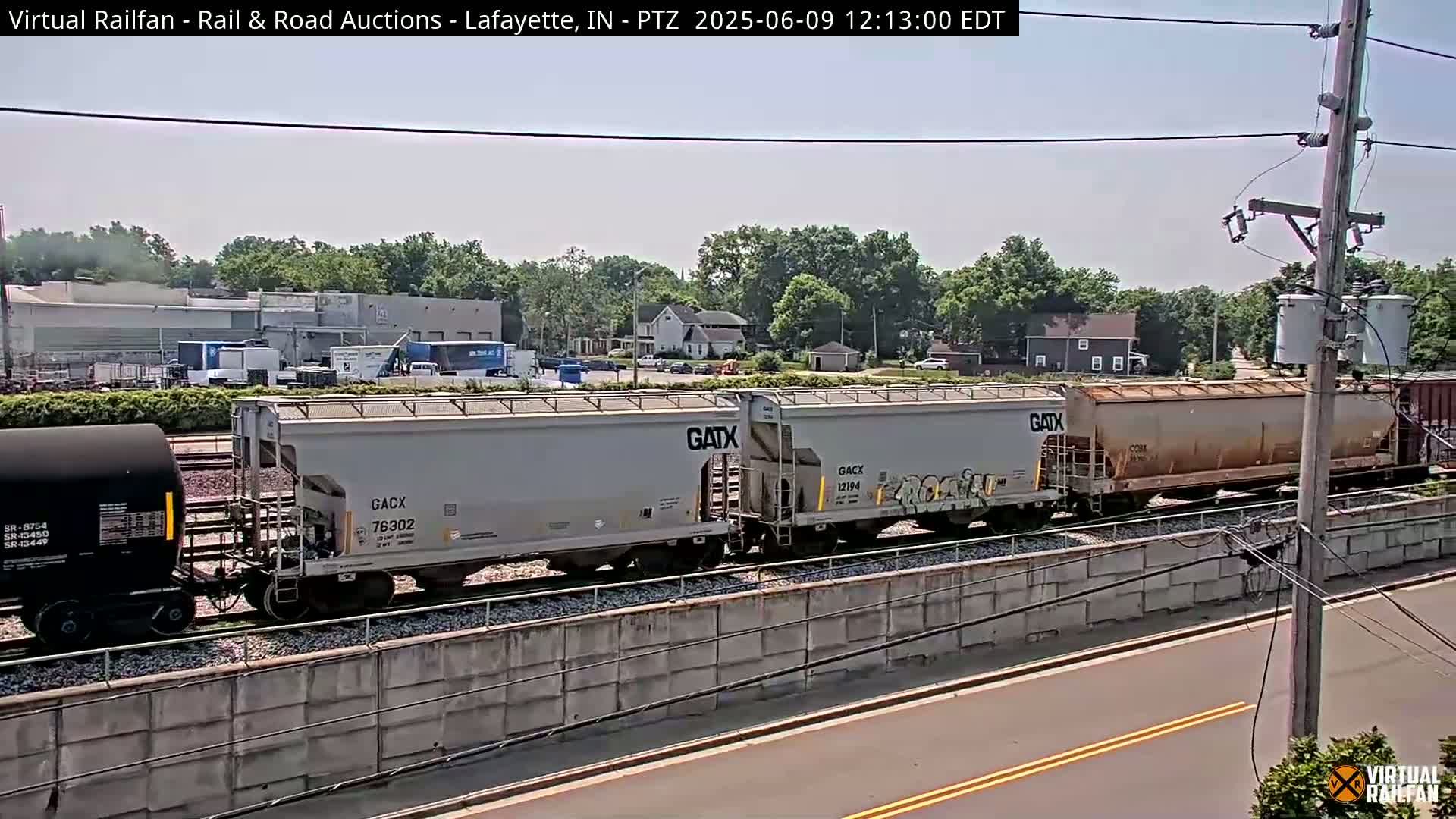 A freight train, consisting of several hopper cars and a tank car, is seen traveling on a railway line under a mostly sunny sky, with some trees and buildings visible in the background.