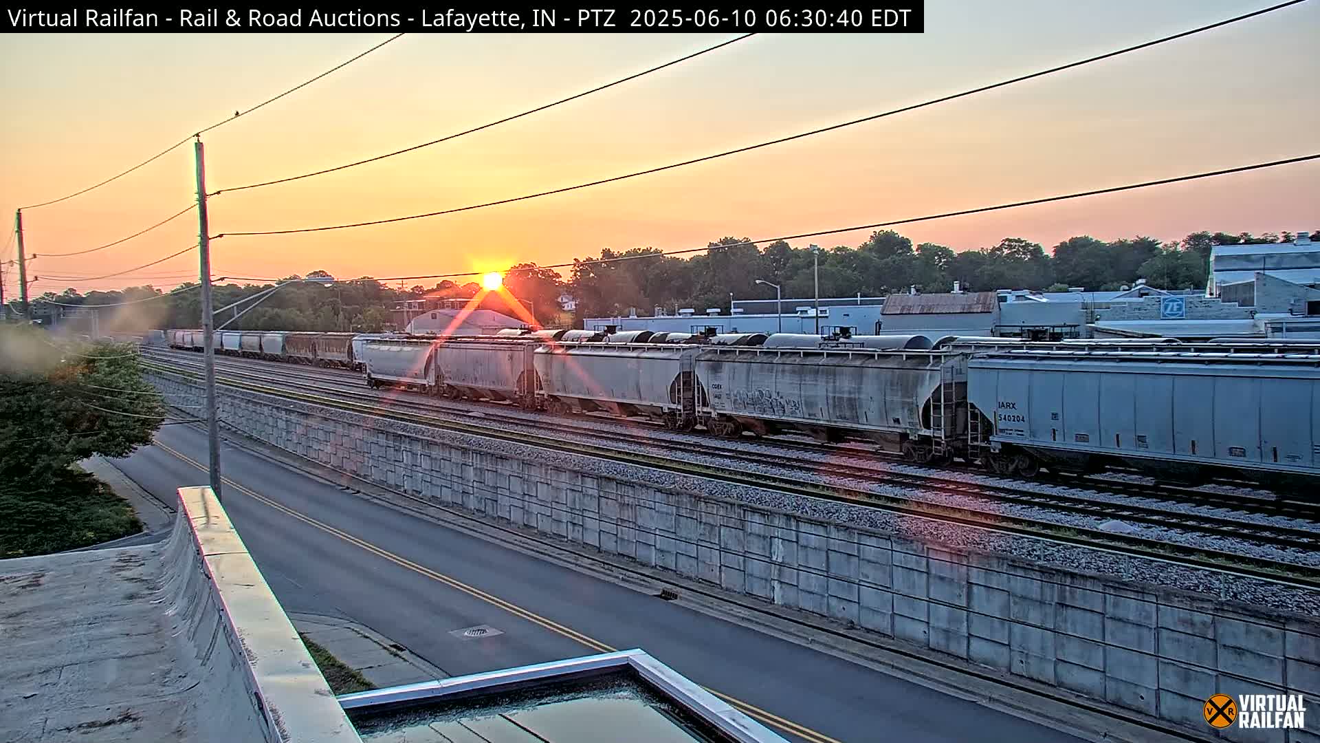 A long freight train passes by at sunset, next to a road and a concrete wall.