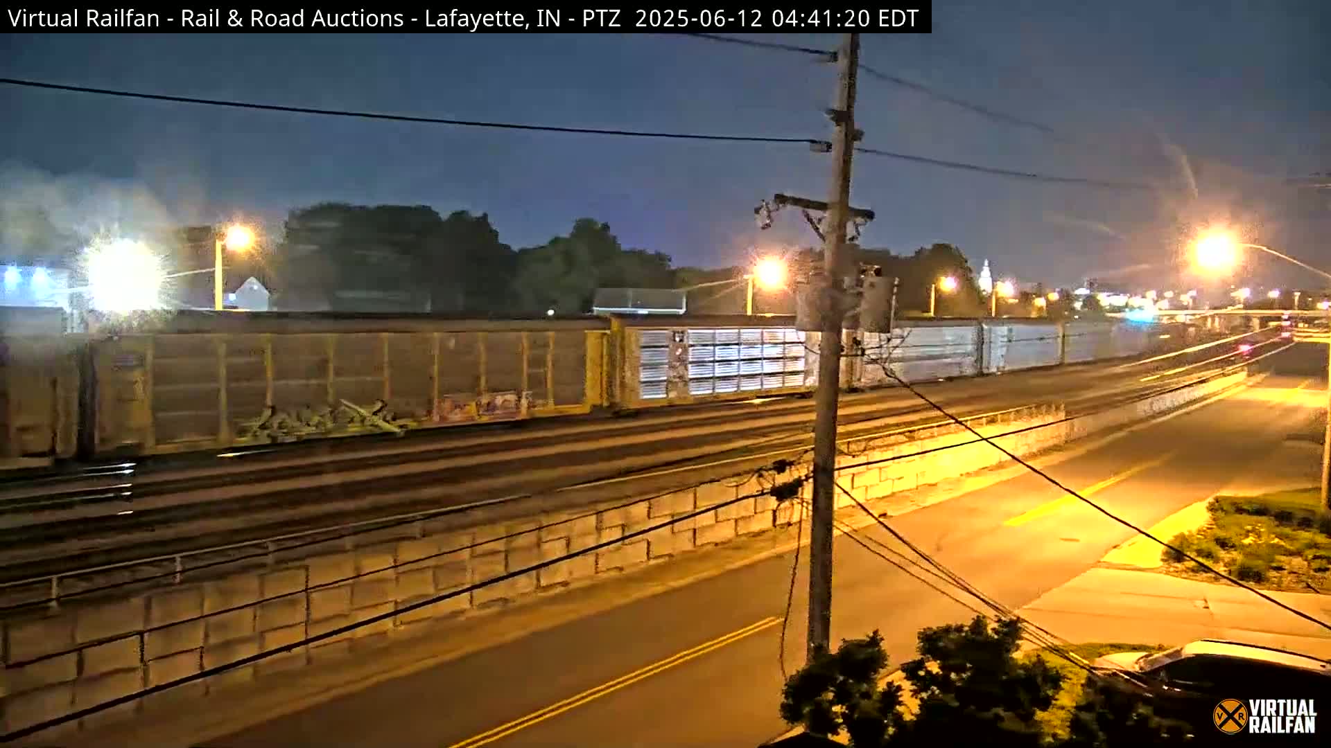 A long freight train passes at night along railroad tracks next to a street, illuminated by streetlights.