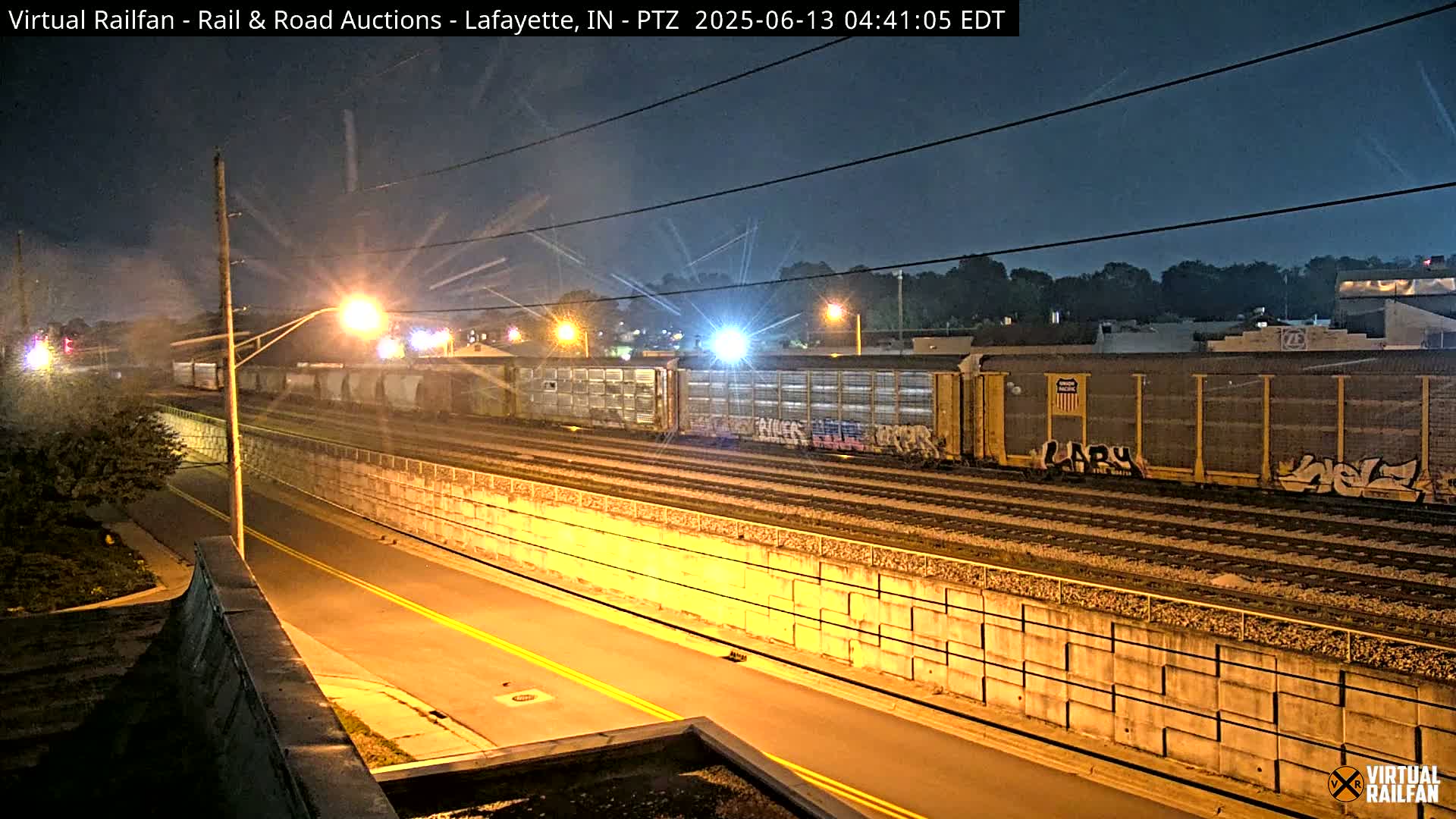 At night, a freight train passes under streetlights next to a roadway separated by a concrete retaining wall.