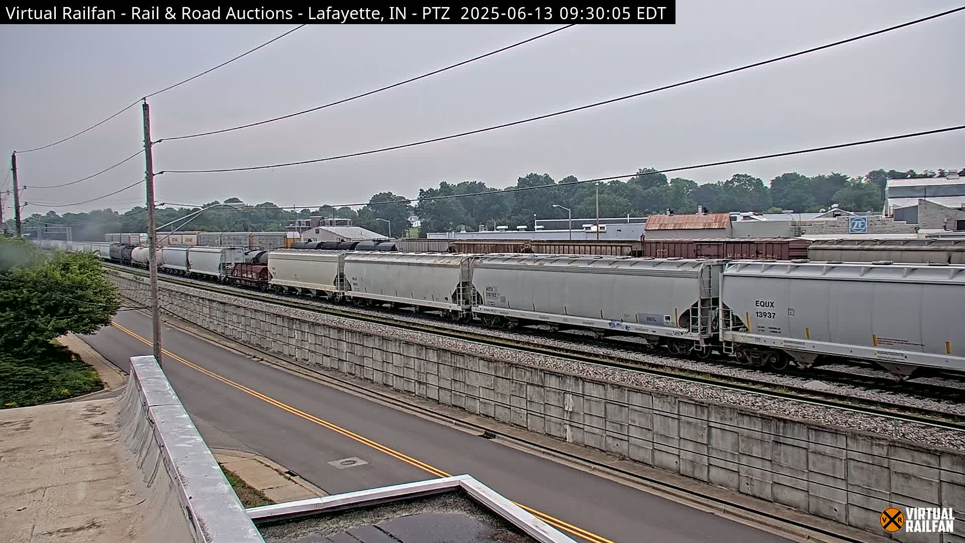 A long freight train travels along railway tracks next to a road under an overcast sky.