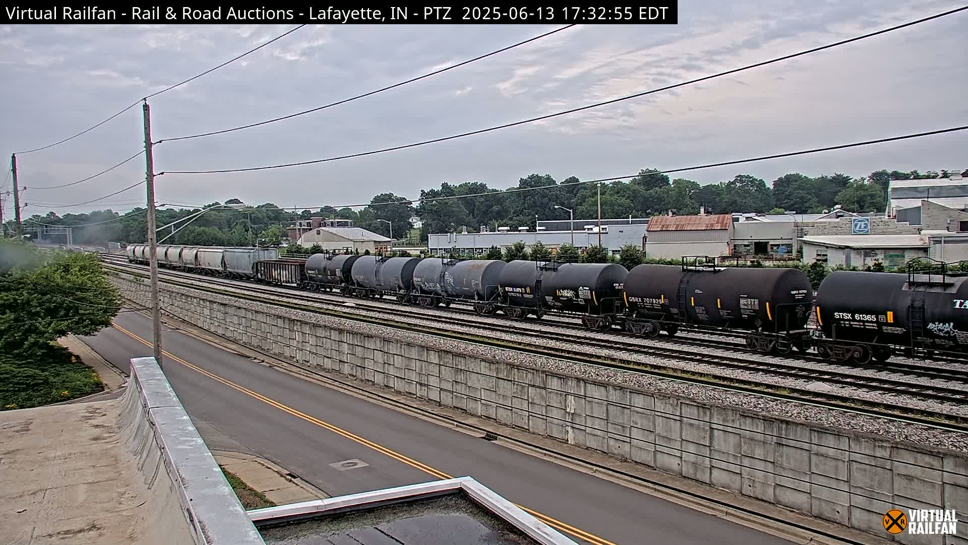 A long freight train, consisting of both covered and tanker cars, moves along a railway track beside a road under an overcast sky.
