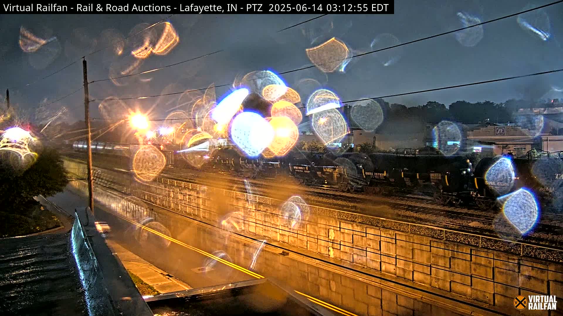 A nighttime view of a rain-soaked train passing by a wet roadway, seen through a rain-covered window.