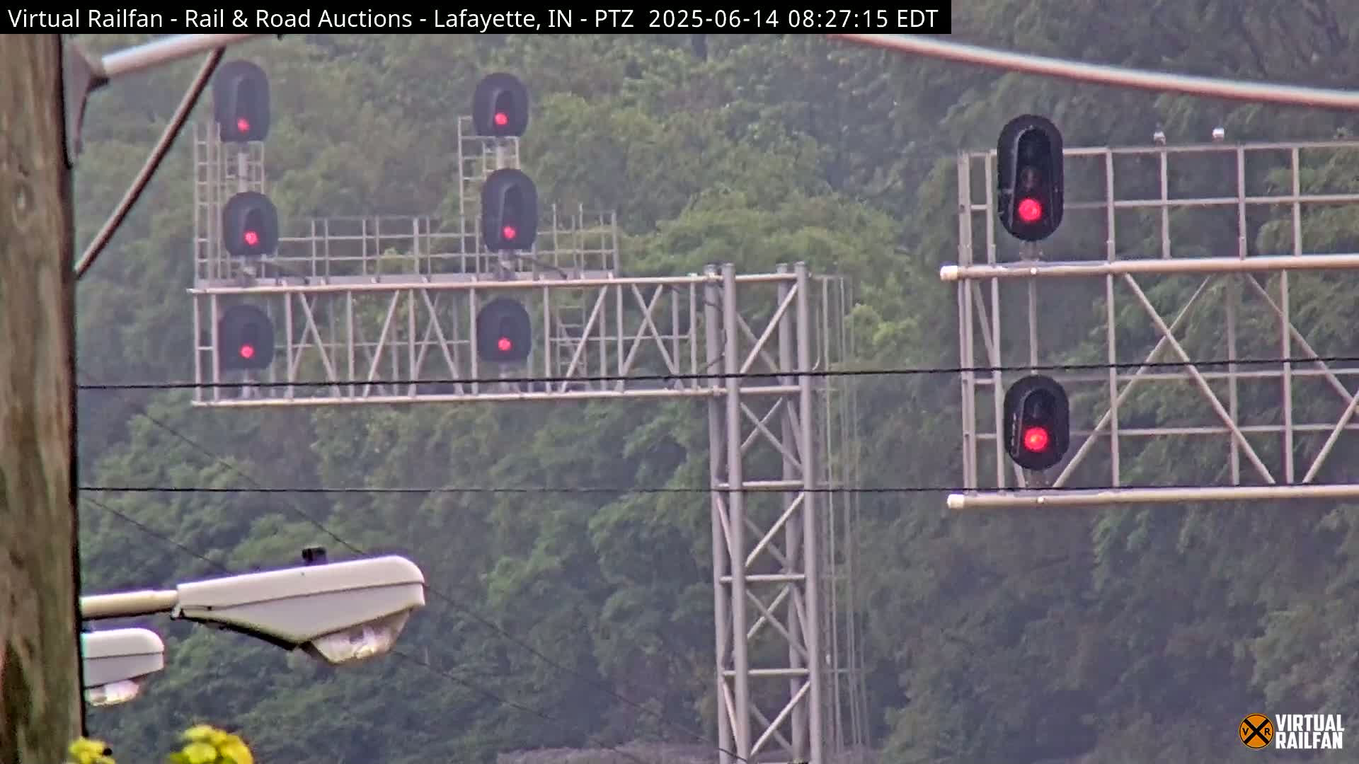 A hazy view of several red railroad signal lights on tall metal structures against a backdrop of green trees.