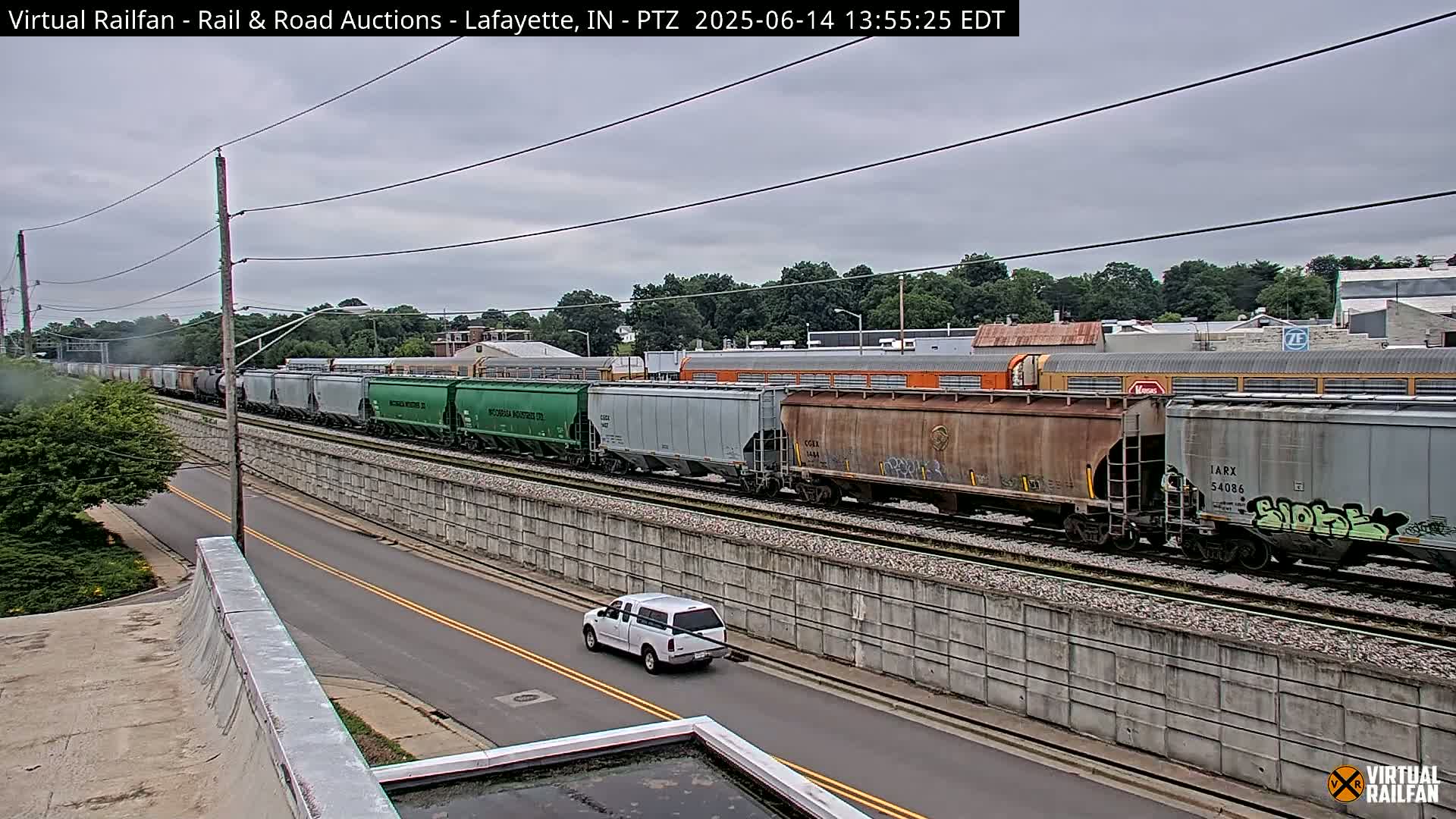 A long freight train passes by a road under an overcast sky, with a white pickup truck driving alongside the tracks.