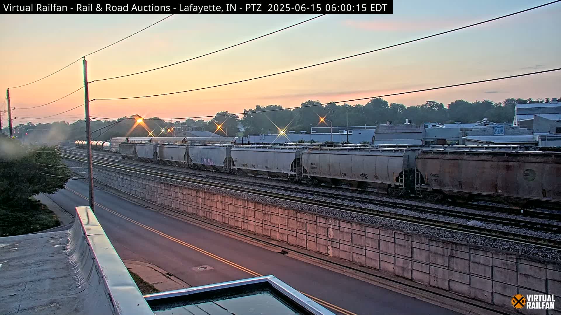 A long freight train passes by under a sunrise sky next to a road and a concrete retaining wall.