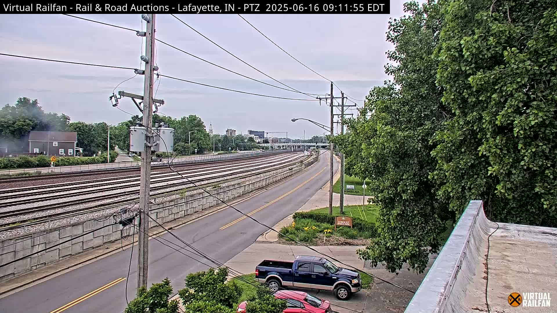 A mostly cloudy day reveals multiple sets of train tracks alongside a two-lane road, bordered by green trees and a pickup truck parked nearby.