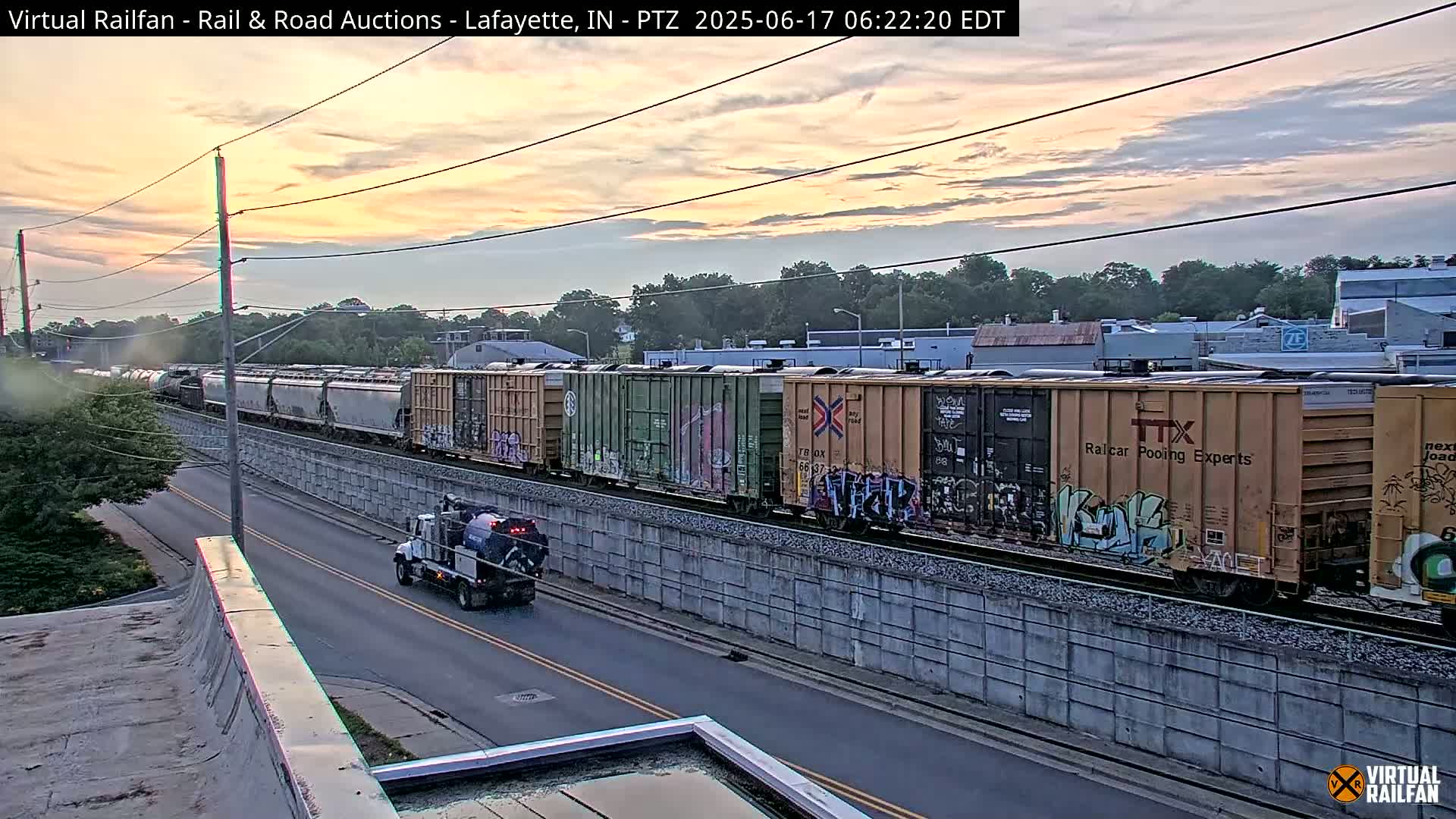 Under a twilight sky, a long freight train passes by a road, with a small service truck driving alongside it.