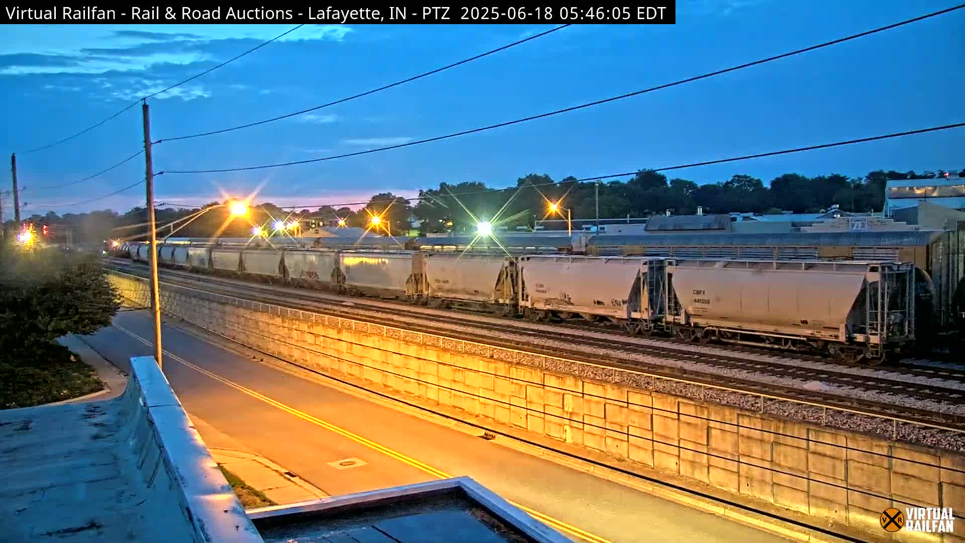 A long freight train of light-grey covered hopper cars sits on a track next to a roadway at twilight under a clear, dark-blue sky.