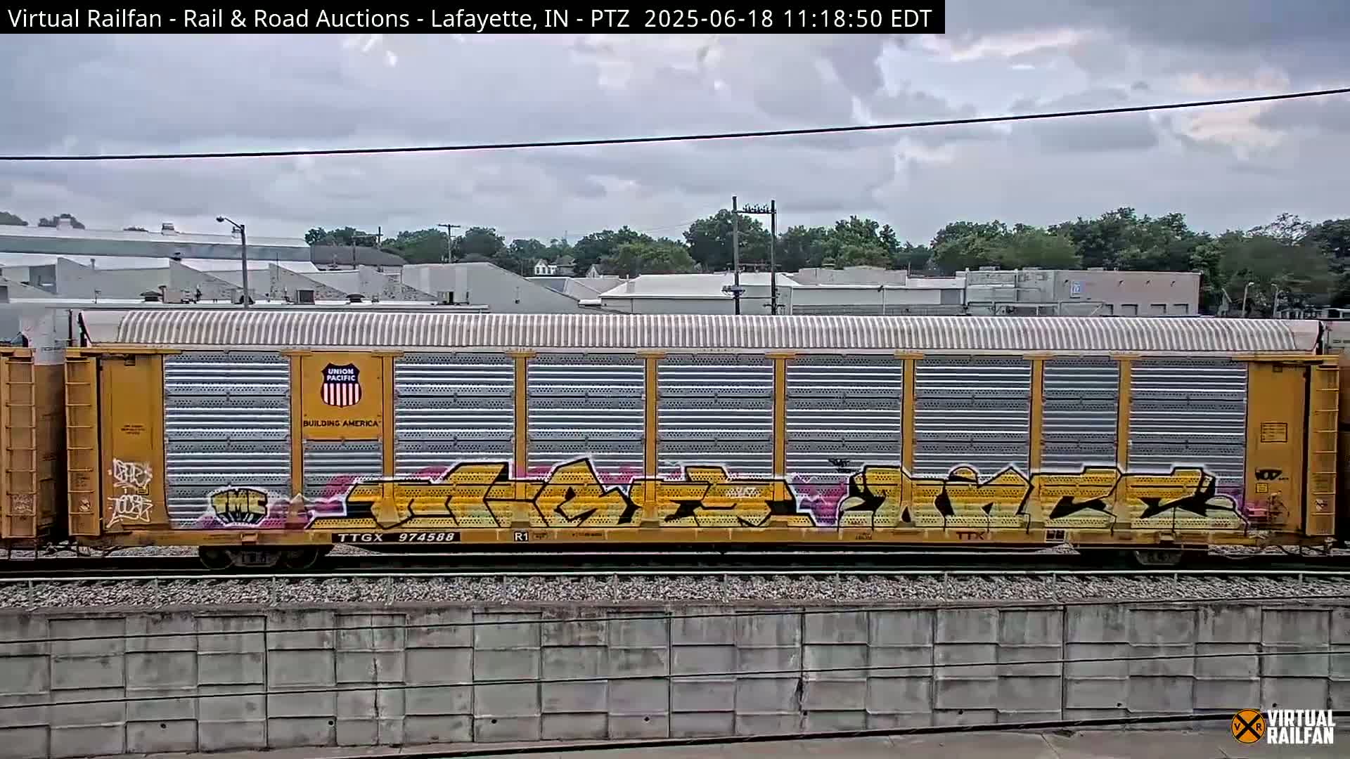 A yellow railroad car with graffiti on its side sits on train tracks under a cloudy sky.