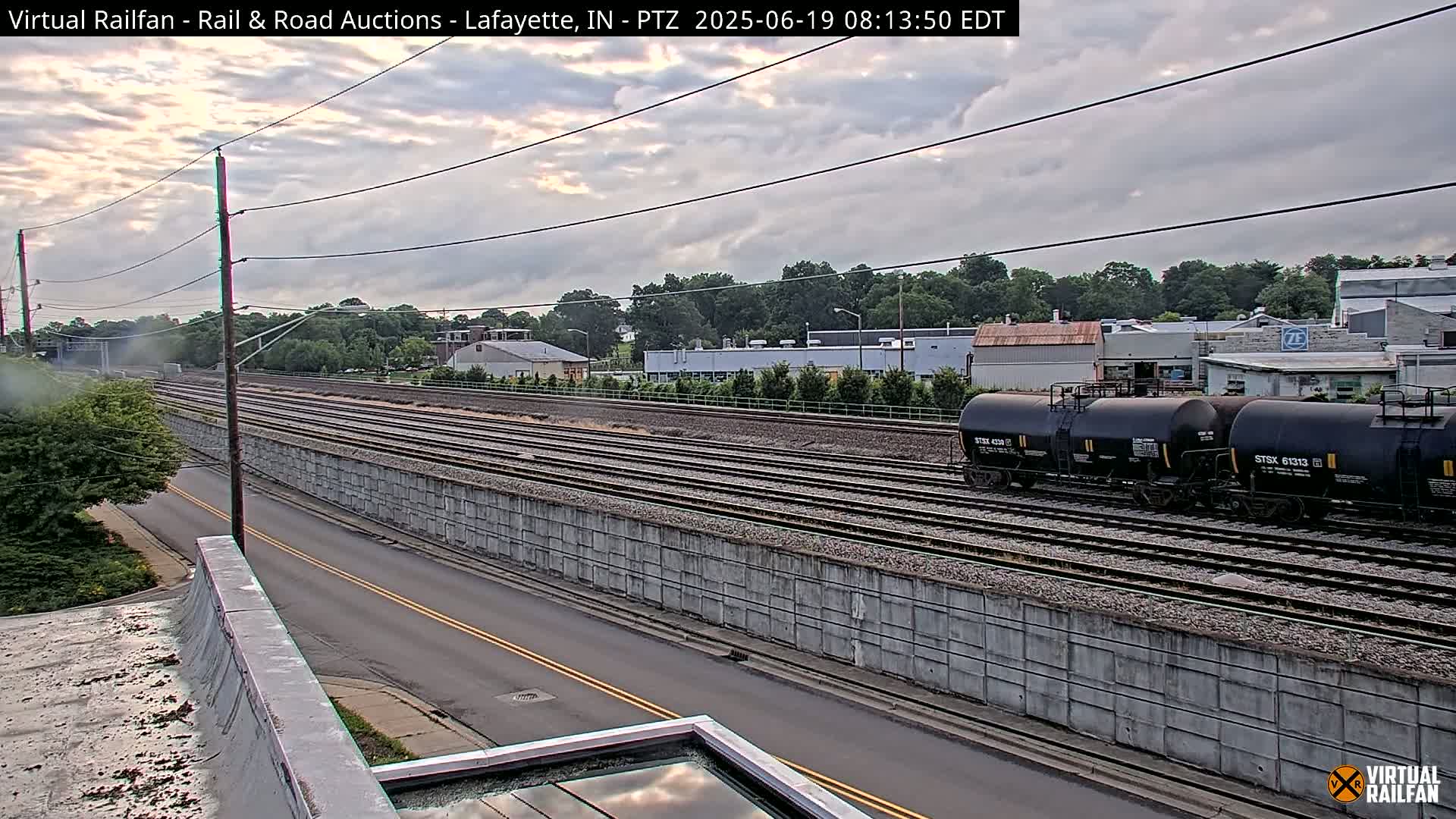 A partly cloudy sky overlooks a railroad track with several tanker cars and a parallel road, bordered by a concrete sound barrier.