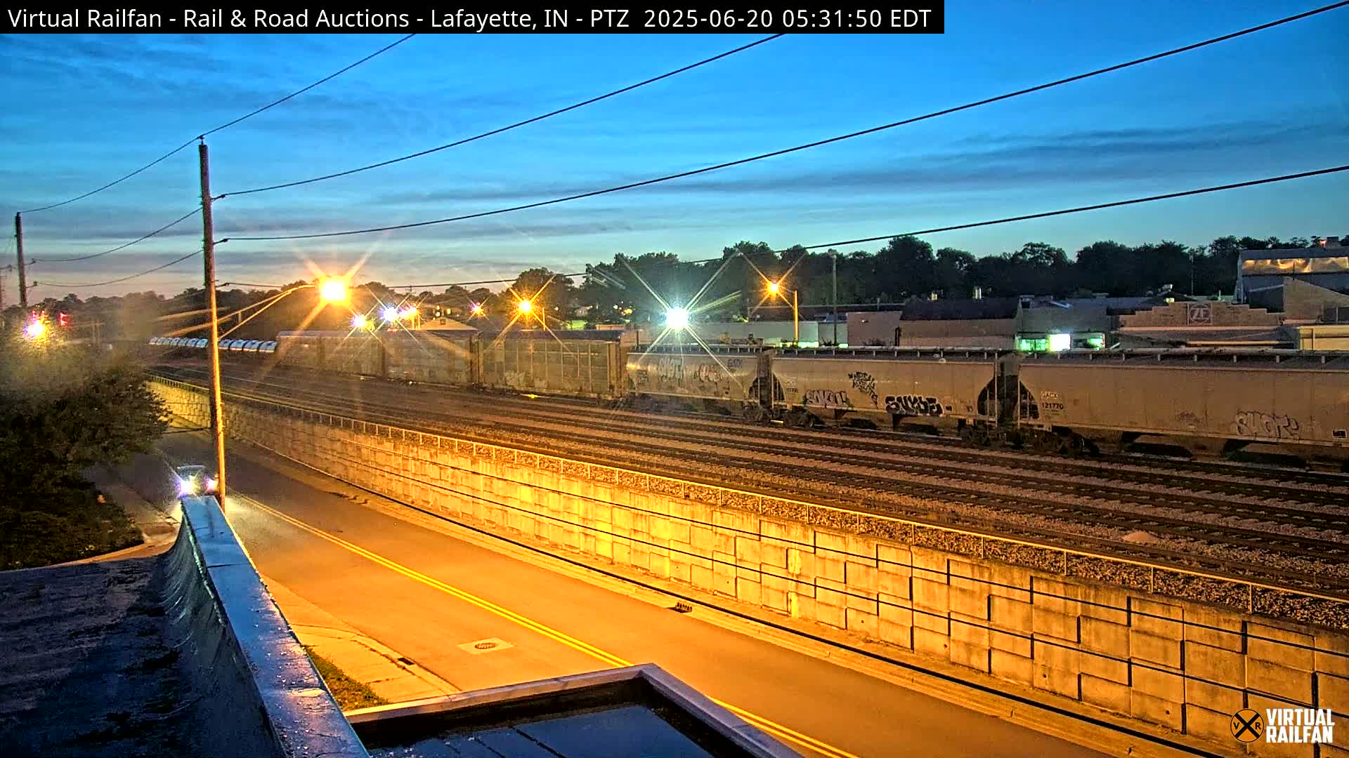 Under a twilight sky, a freight train sits on tracks beside a roadway separated by a concrete retaining wall.