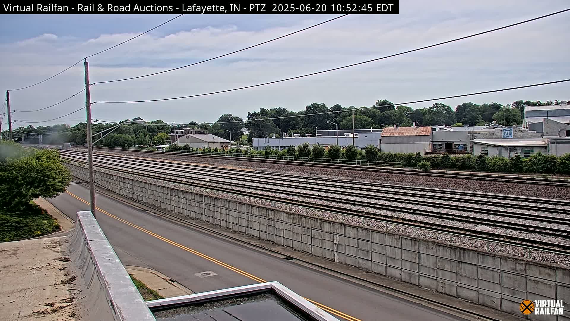 A mostly sunny day shows a view of multiple train tracks next to a road, all bordered by a tall concrete wall, with trees and buildings in the background.