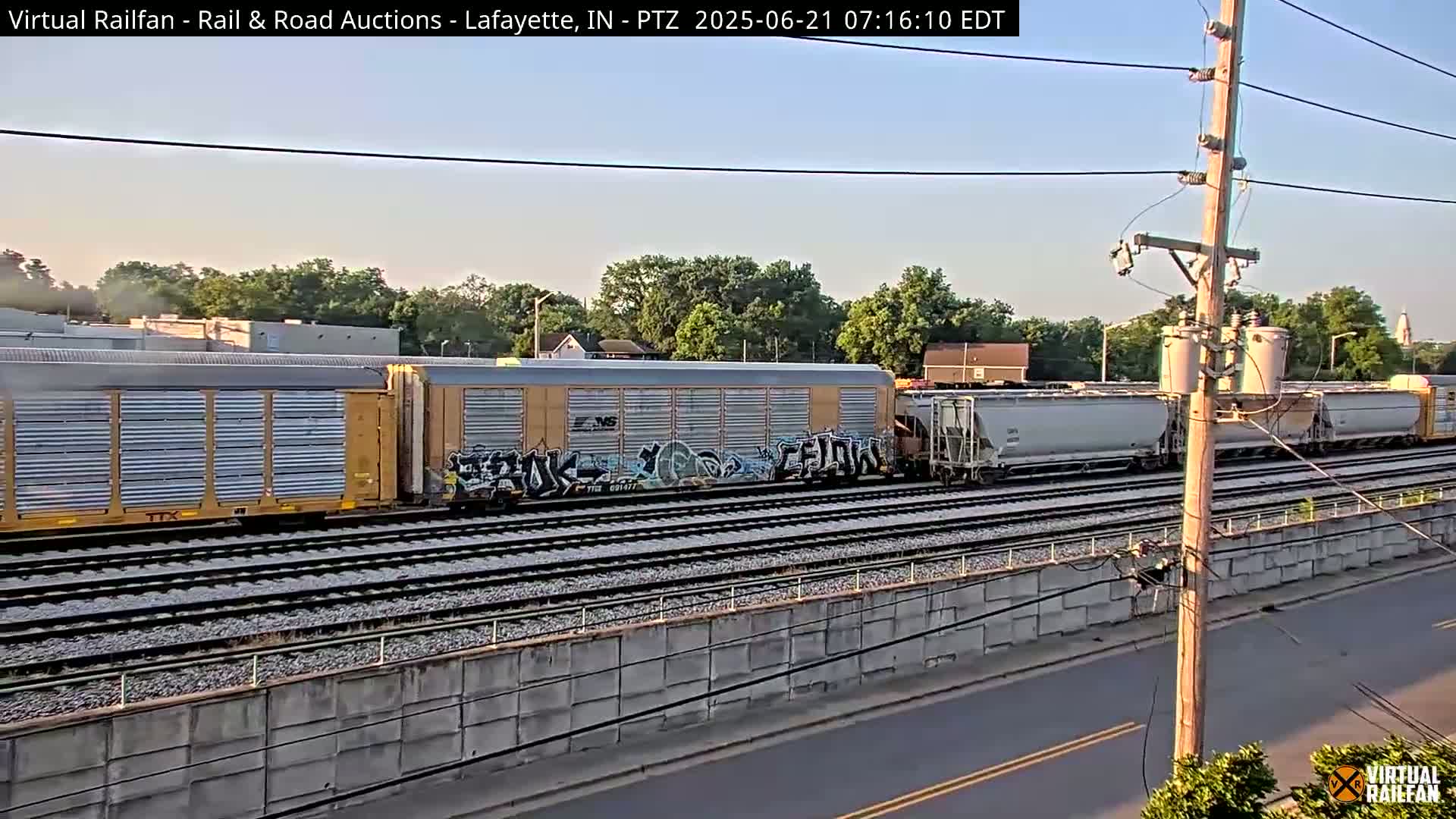 A long freight train with graffiti on some cars passes by under a mostly clear sky.