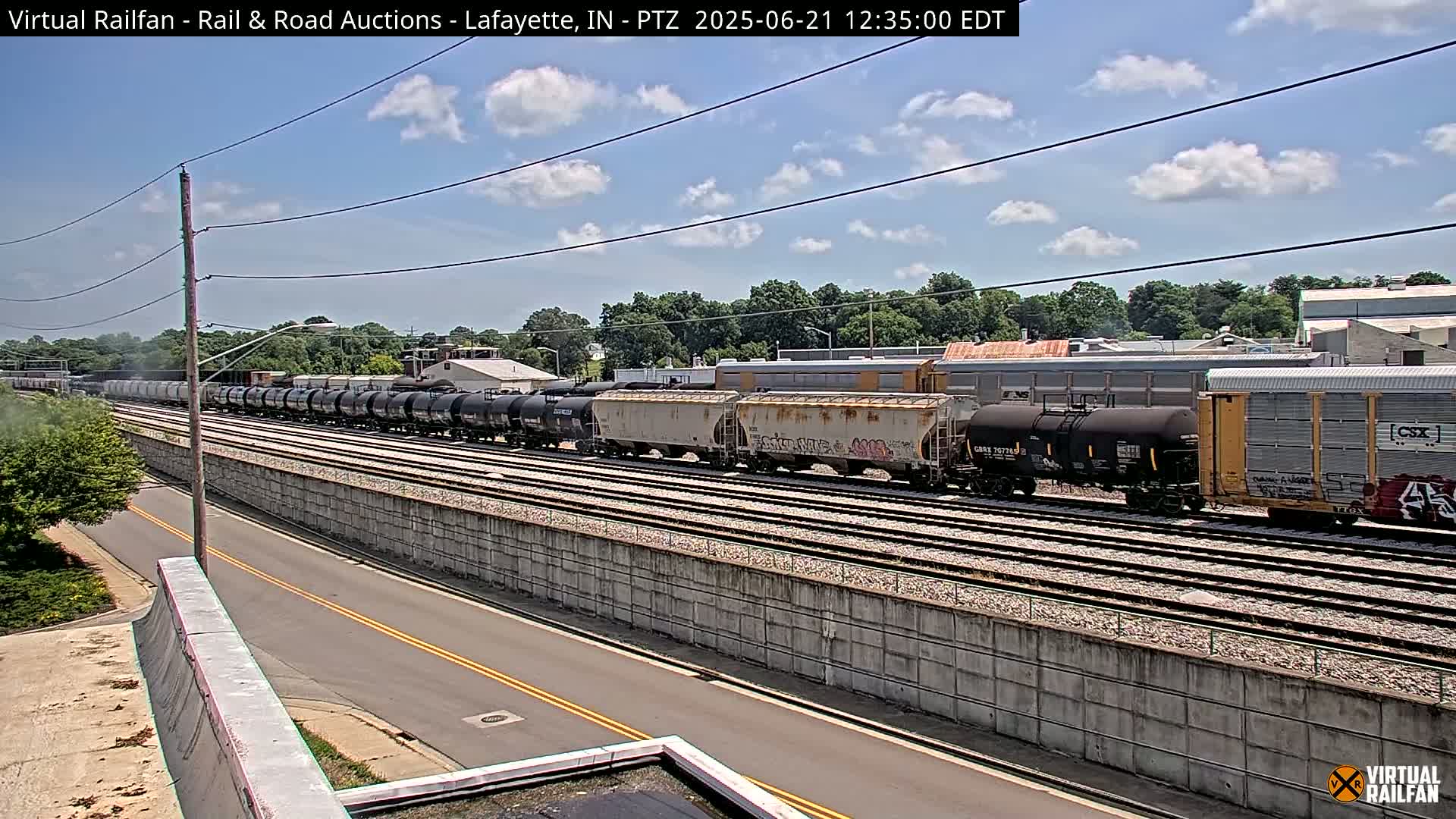 A long freight train of various cars sits on railroad tracks next to a road under a partly cloudy sky.