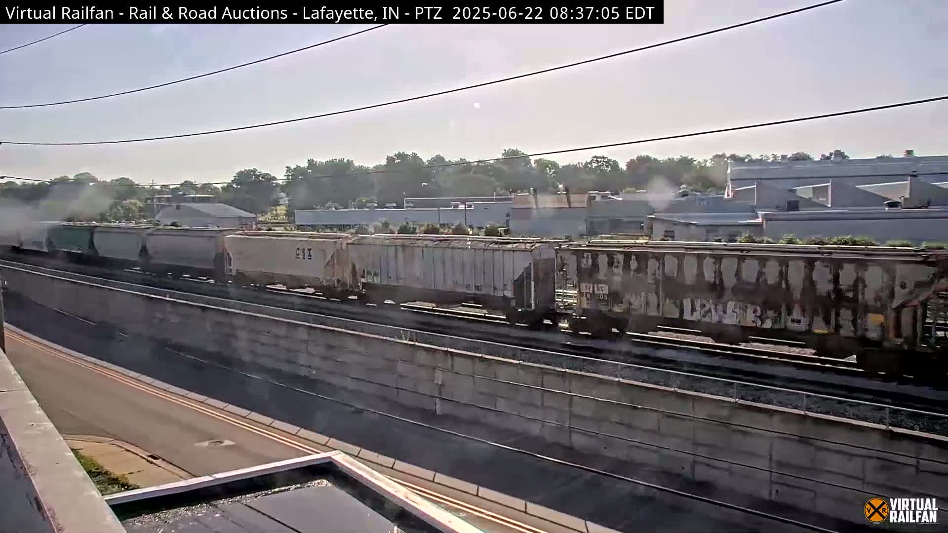 A long freight train passes by on a sunny day, traveling along a railway next to a road and a concrete wall.
