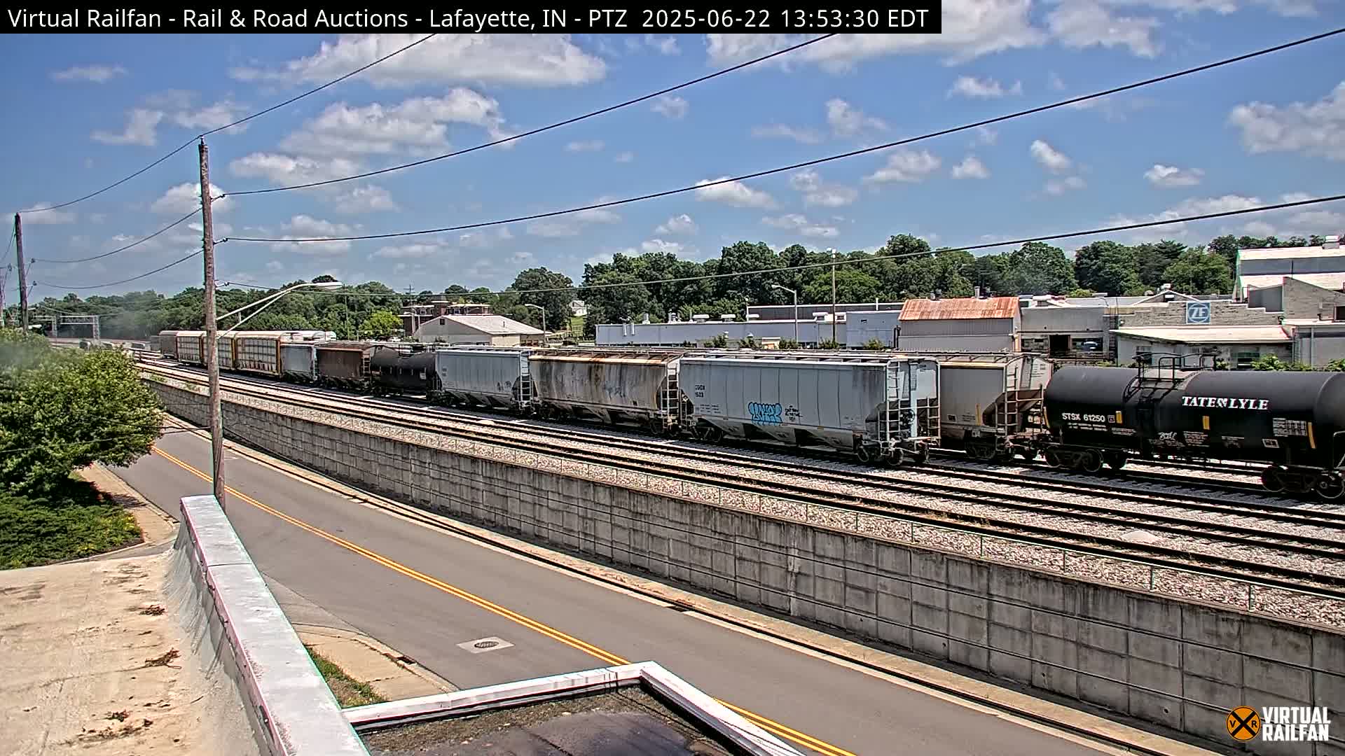 A long freight train passes by on a sunny day, next to a road bordered by a concrete wall.