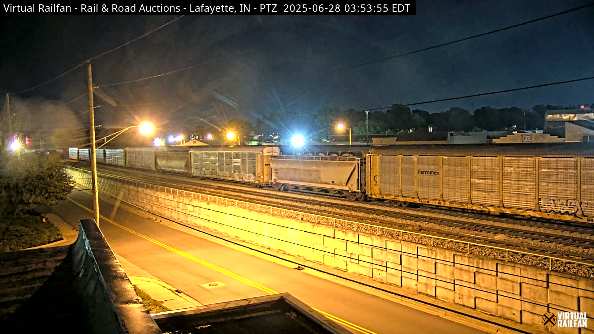 A long freight train moves along railroad tracks at night, next to a roadway separated by a concrete wall under a dark, clear sky.