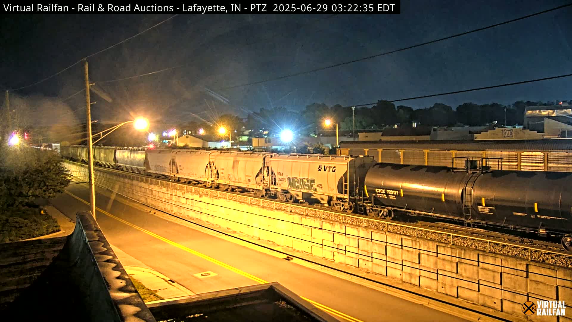 A freight train, illuminated by streetlights at night, passes along a raised section of track beside a road.