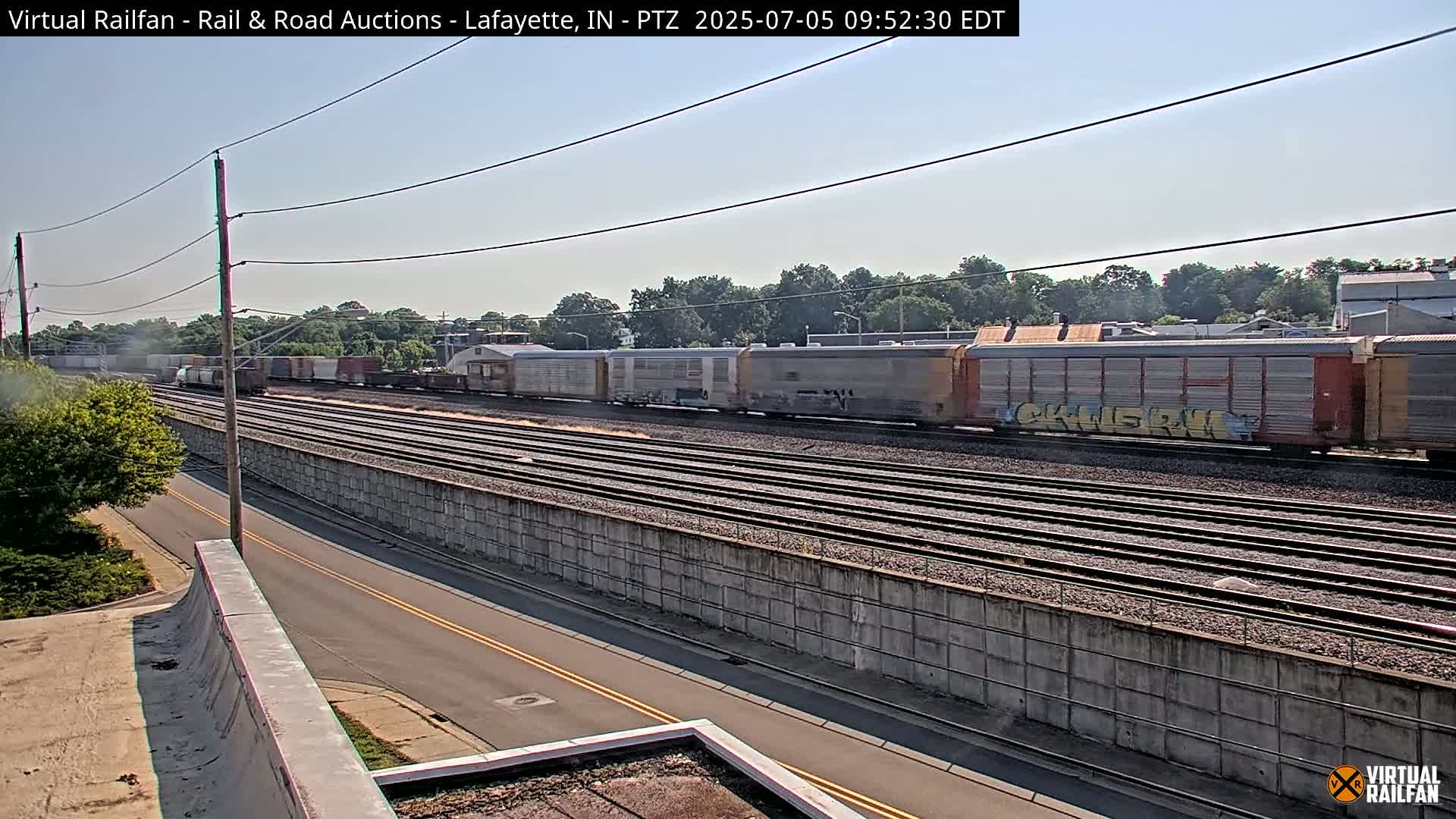 A long freight train sits on multiple parallel train tracks under a clear, sunny sky, next to a road and a concrete sound barrier.