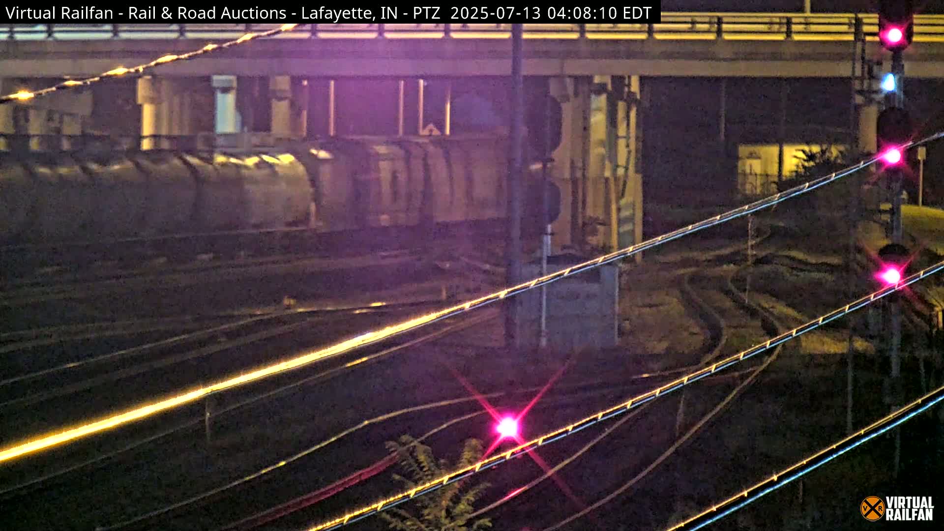 At night, a long freight train sits under a bridge near some railroad tracks and signal lights.