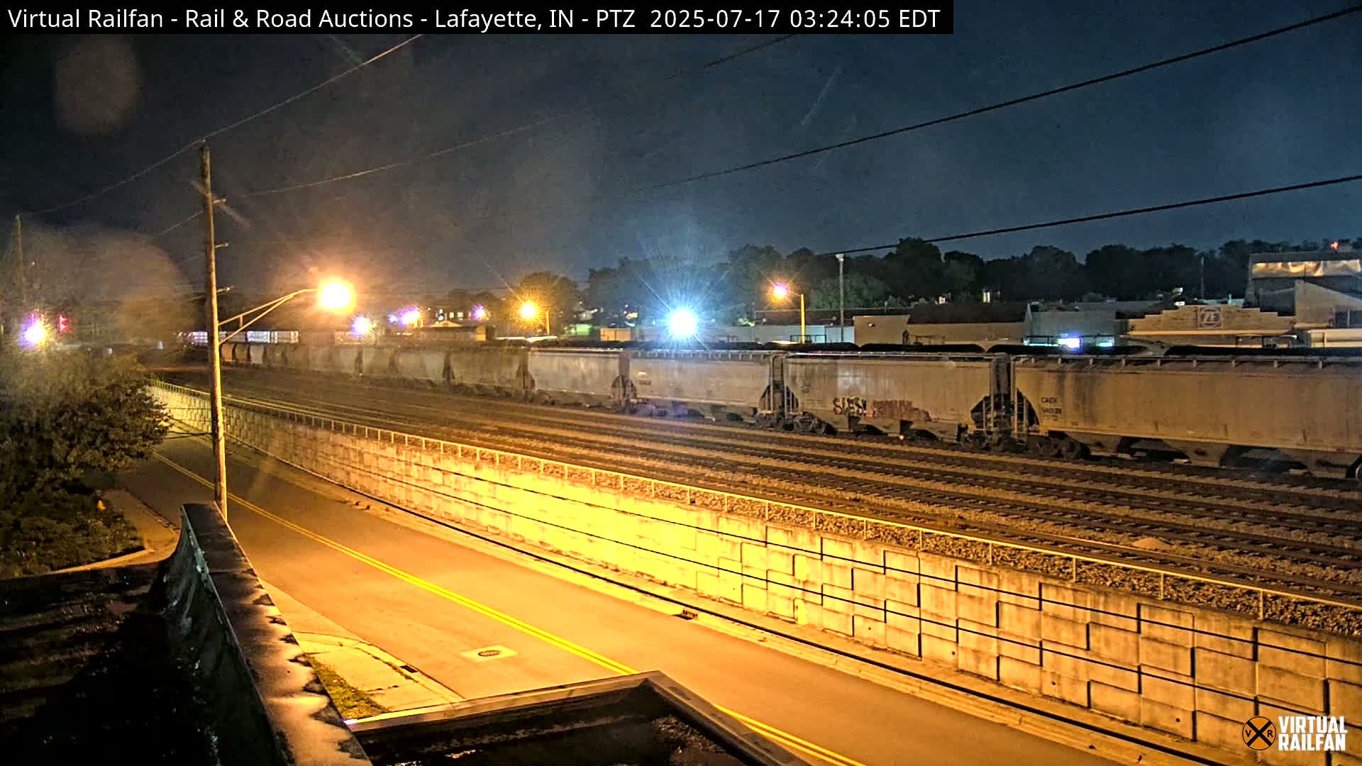At night, a train of boxcars sits on railroad tracks next to a roadway, separated by a concrete barrier.