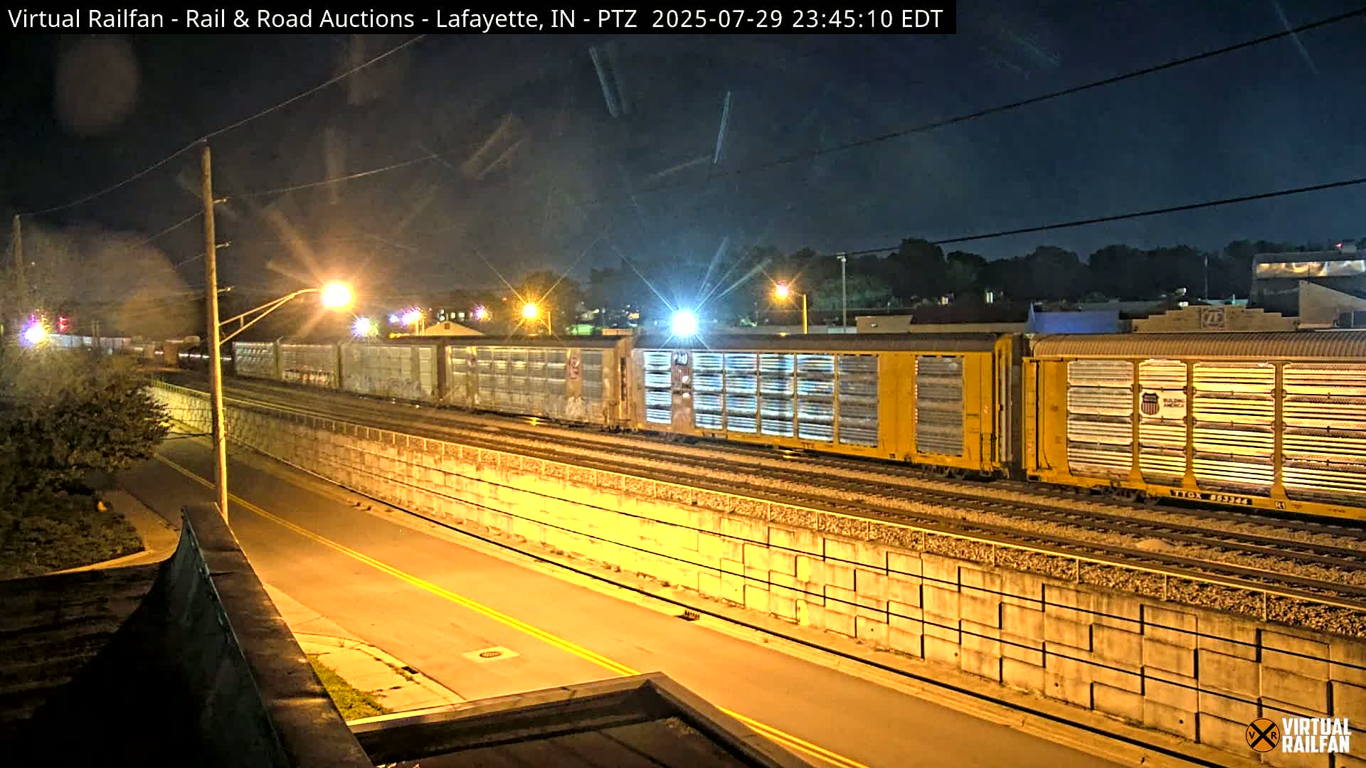 A nighttime view of railroad tracks under a bridge, illuminated by lights and possibly rain.