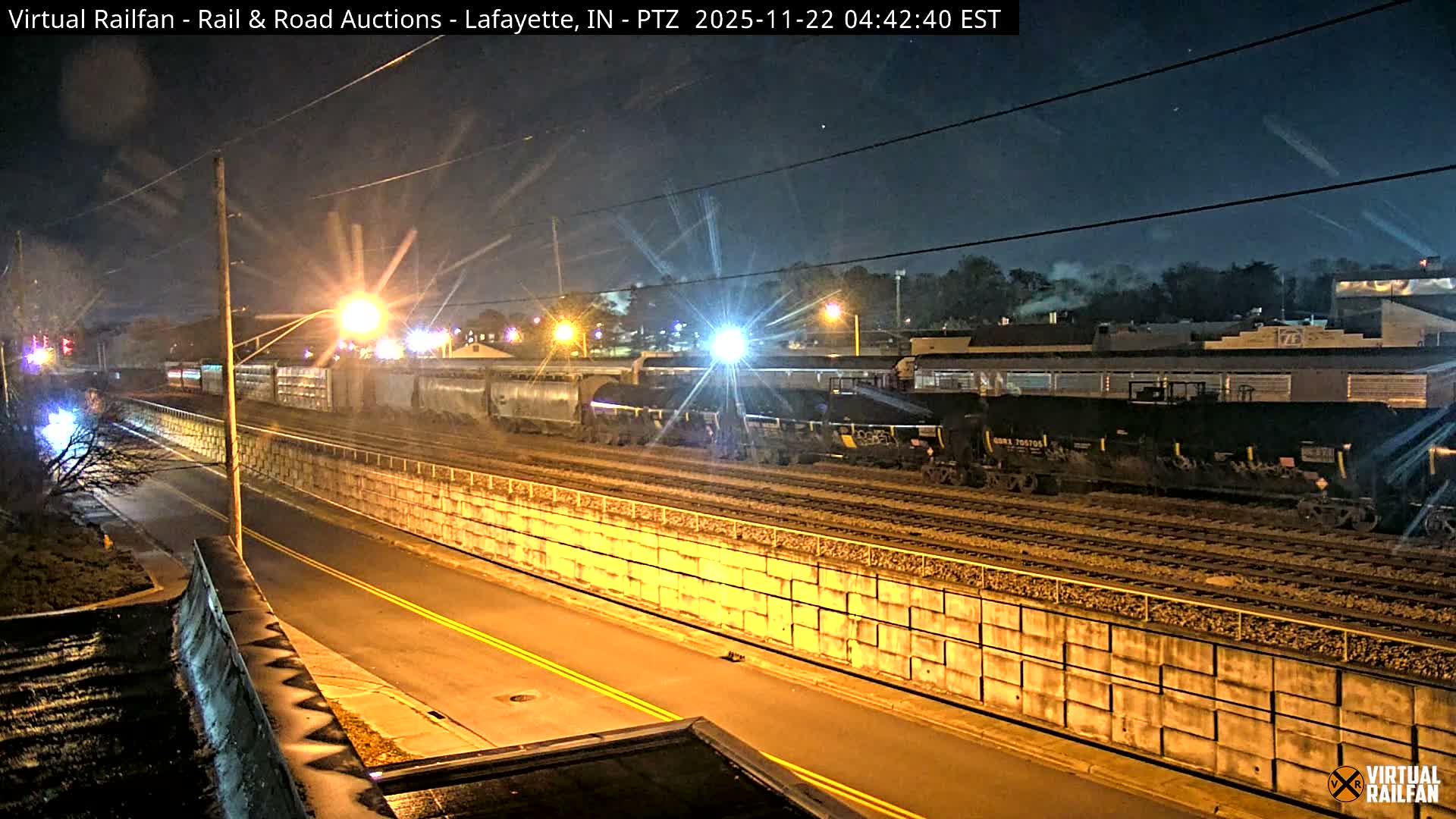 Under a dark, misty night sky, a bright headlight leads a freight train on multiple tracks next to a wet road illuminated by streetlights, with various industrial buildings in the background.