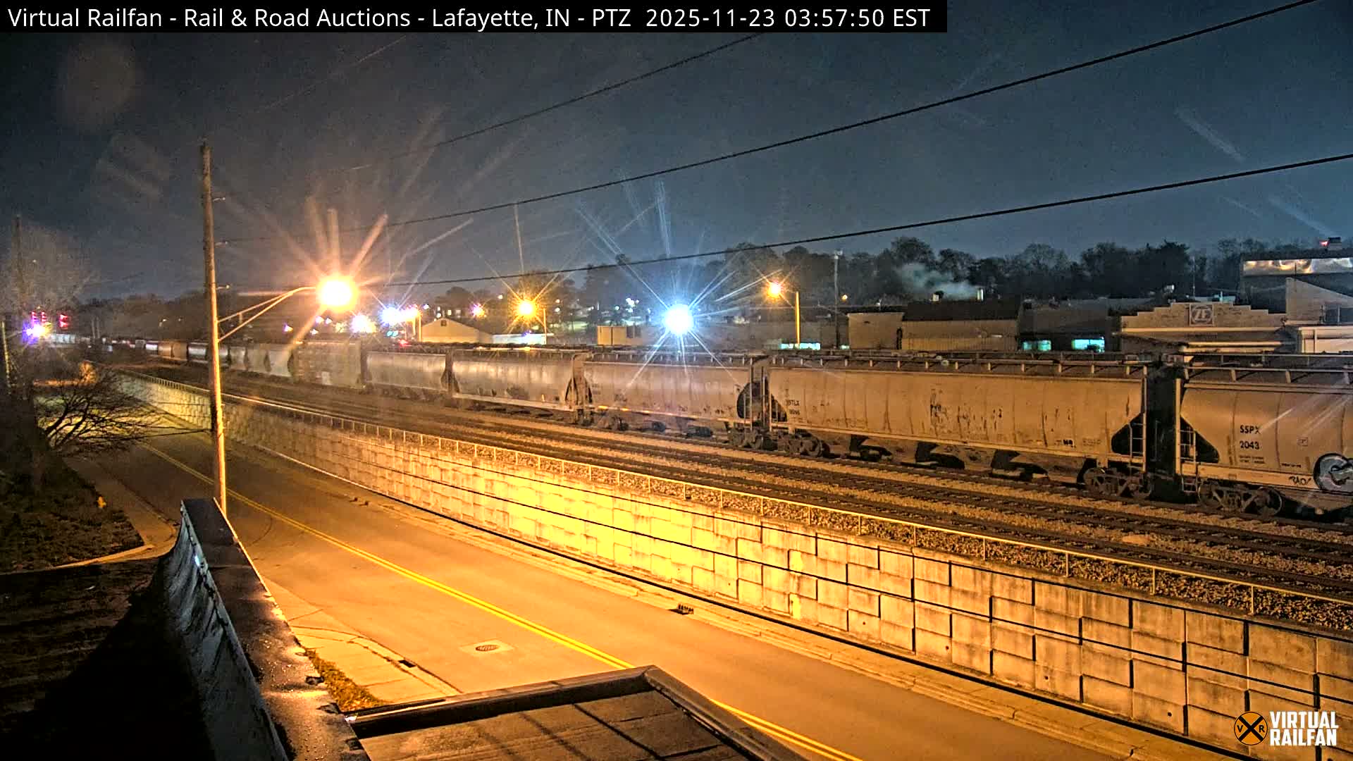 A lengthy freight train occupies multiple tracks alongside a brightly lit street at night, with buildings and power lines visible in the distance under a clear, dark sky.
