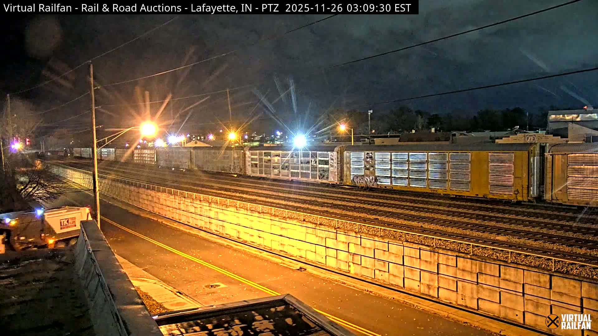 An illuminated train with several yellow and silver cars sits on multiple tracks next to a parallel road with a vehicle, all under a clear night sky, lit by numerous streetlights.